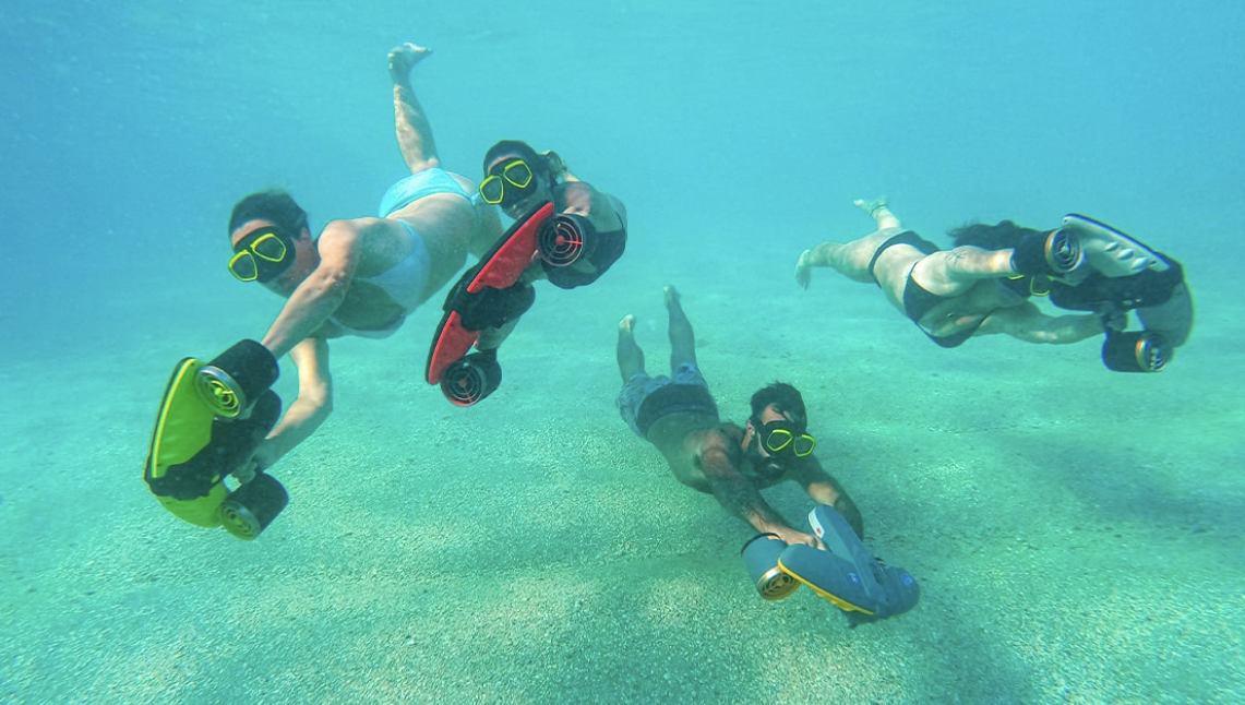 Three people underwater wearing snorkeling masks and fins, holding small underwater scooters, floating above a sandy ocean floor.