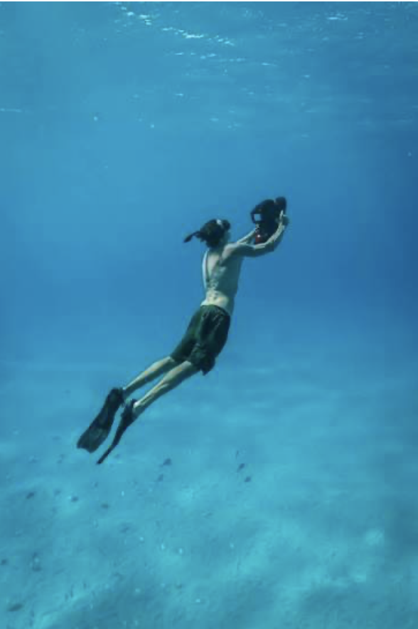 A person underwater holding a camera with both hands, wearing swim fins and dark swim trunks, with the ocean floor visible below.