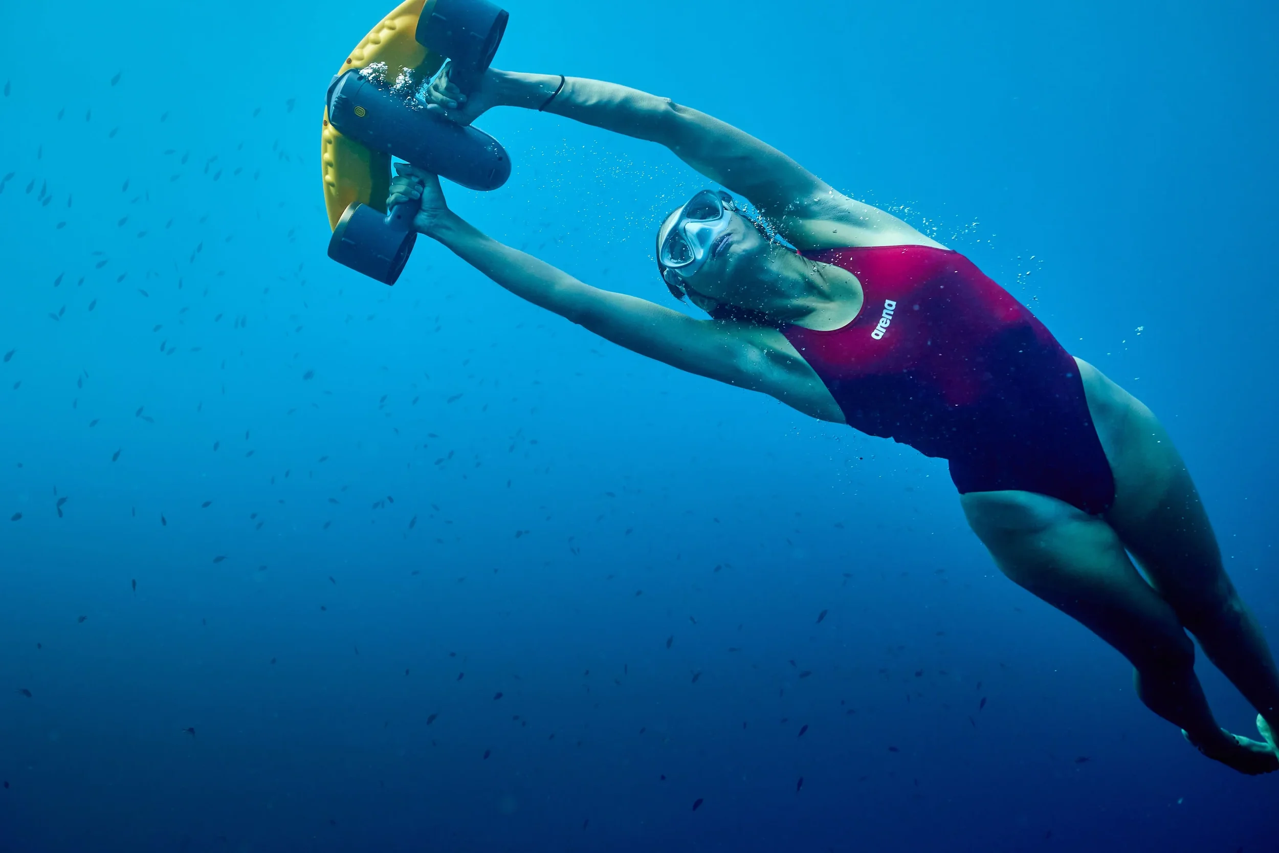 A woman holding a yellow sea scooter under water in a red swimsuit with goggles