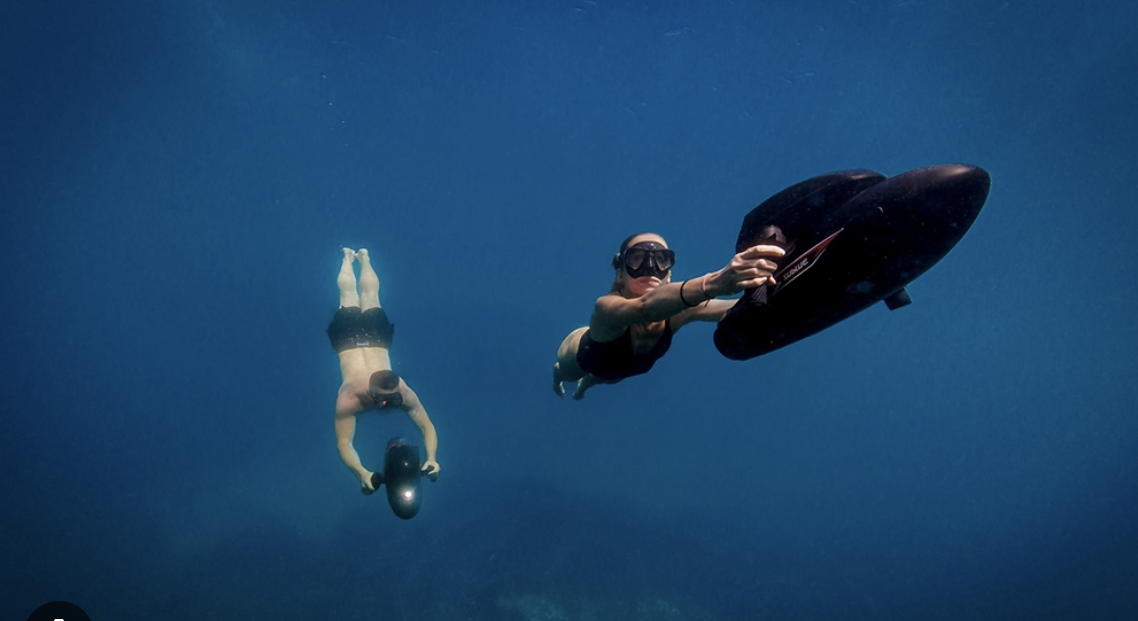 Two people scuba diving underwater; one woman holding a large black object, the other person is posing downward with their arms extended.