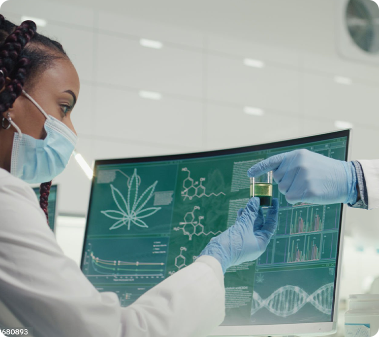 A scientist wearing a face mask and gloves holding a small vial with a green liquid in front of a computer screen displaying chemical diagrams and graphs in a laboratory.