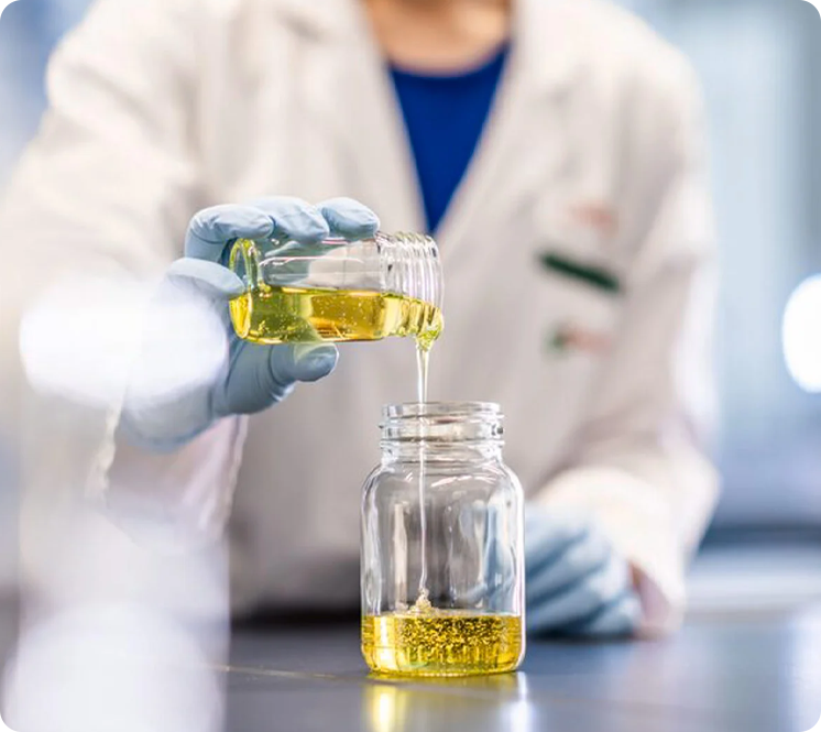 A scientist in a lab coat and gloves pouring yellow liquid from a test tube into a glass jar.
