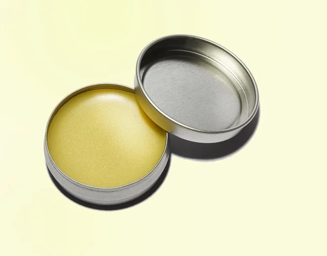 A circular metal tin with yellow lip balm inside and the lid placed next to it on a light surface.