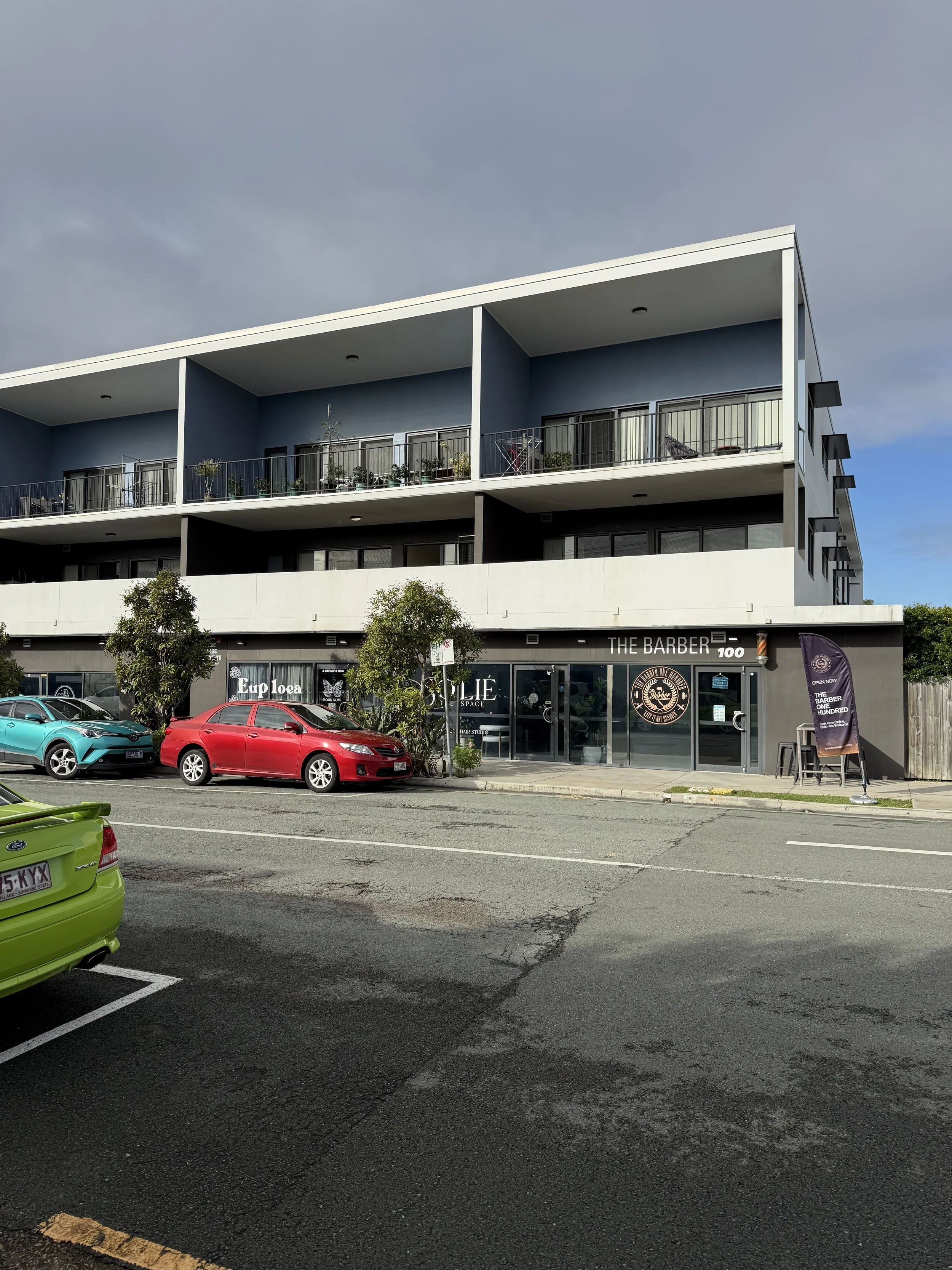 Street view of a three-story building with commercial shops on the ground floor and residential units above, featuring balconies. There are parked cars in the foreground, and trees in front of the building. The sky is cloudy.