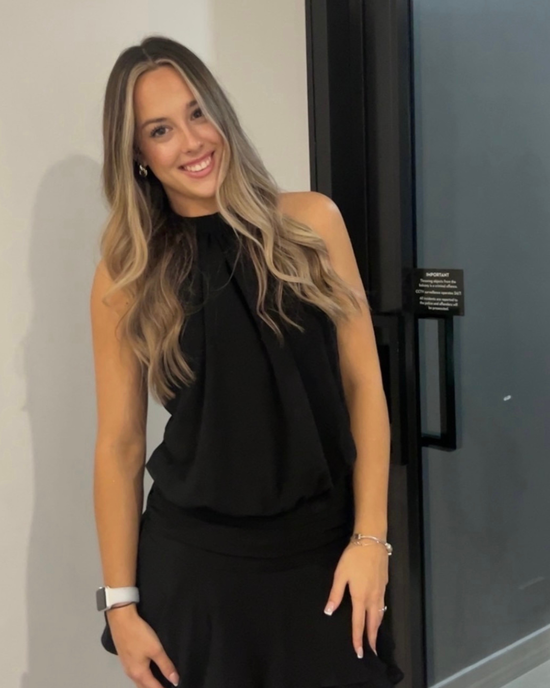 A young woman with long wavy blonde hair, wearing a sleeveless black dress, smiling and posing indoors next to a door.