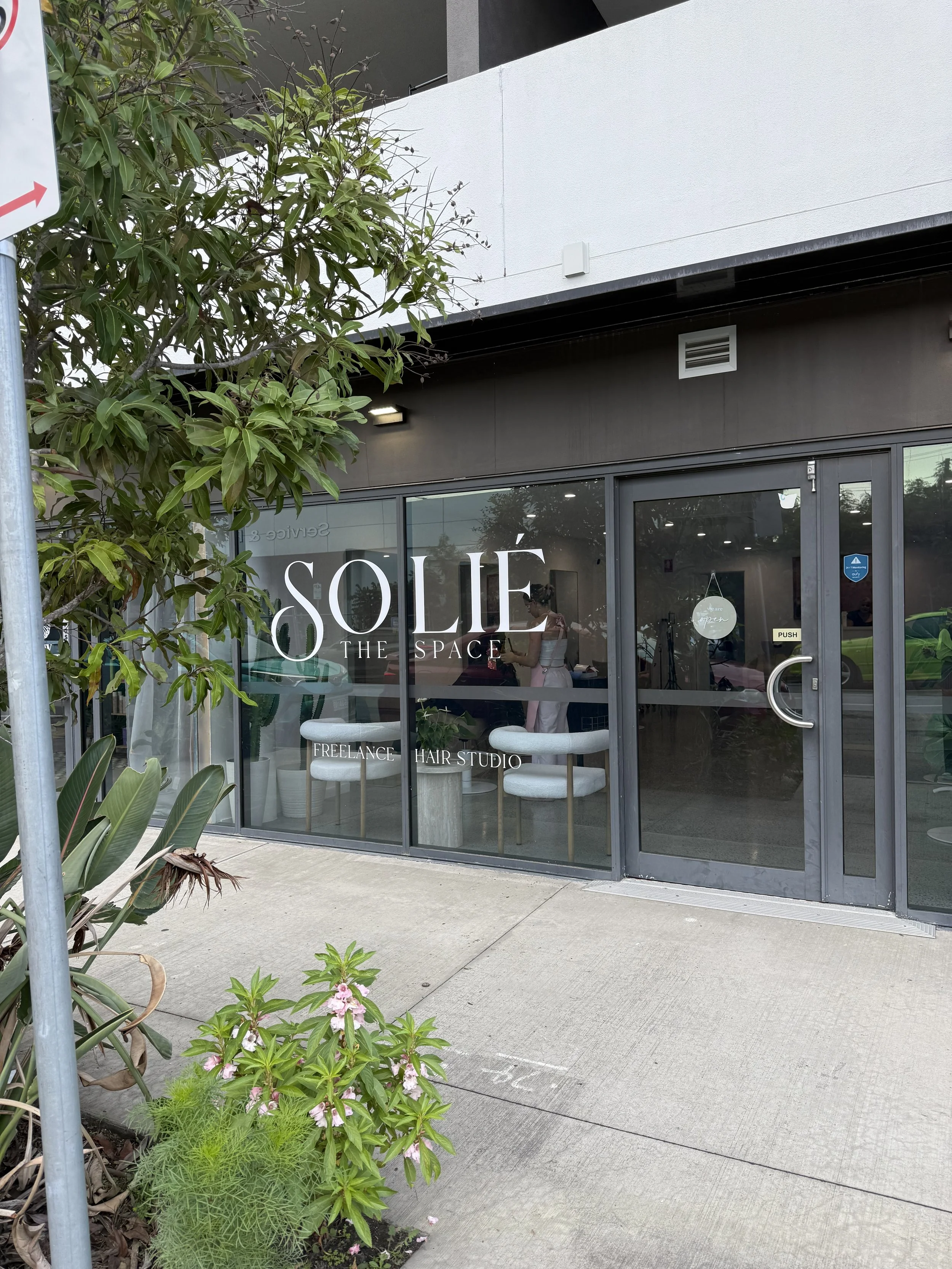 The exterior of a modern hair studio with large glass windows and a sign reading 'SOLLÉ THE SPACE'. There is a small porch with two white chairs inside. Outside, there are various green plants and flowers near the sidewalk.