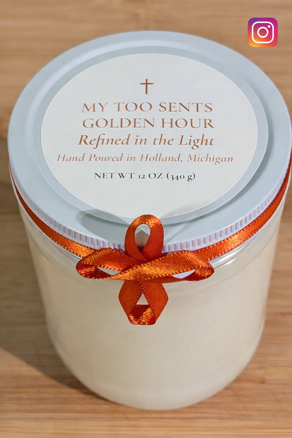 A white candle in a jar with an orange ribbon tied around it, topped with a white lid that has a label reading 'My Too Sents Golden Hour,' handmade in Holland, Michigan.