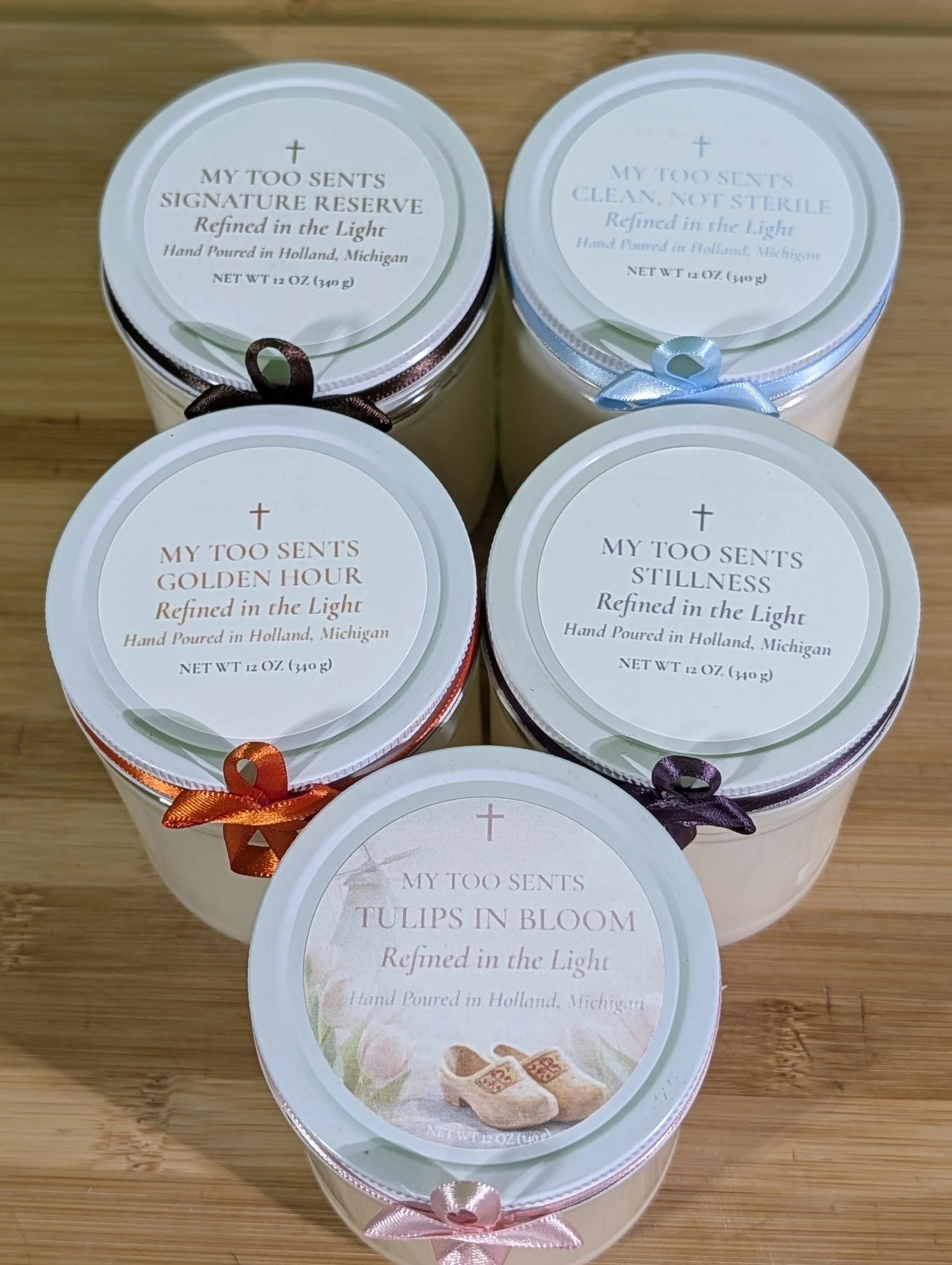 Five scented candles in white containers with different colored ribbons on a wooden surface, labeled with various scents and descriptions.
