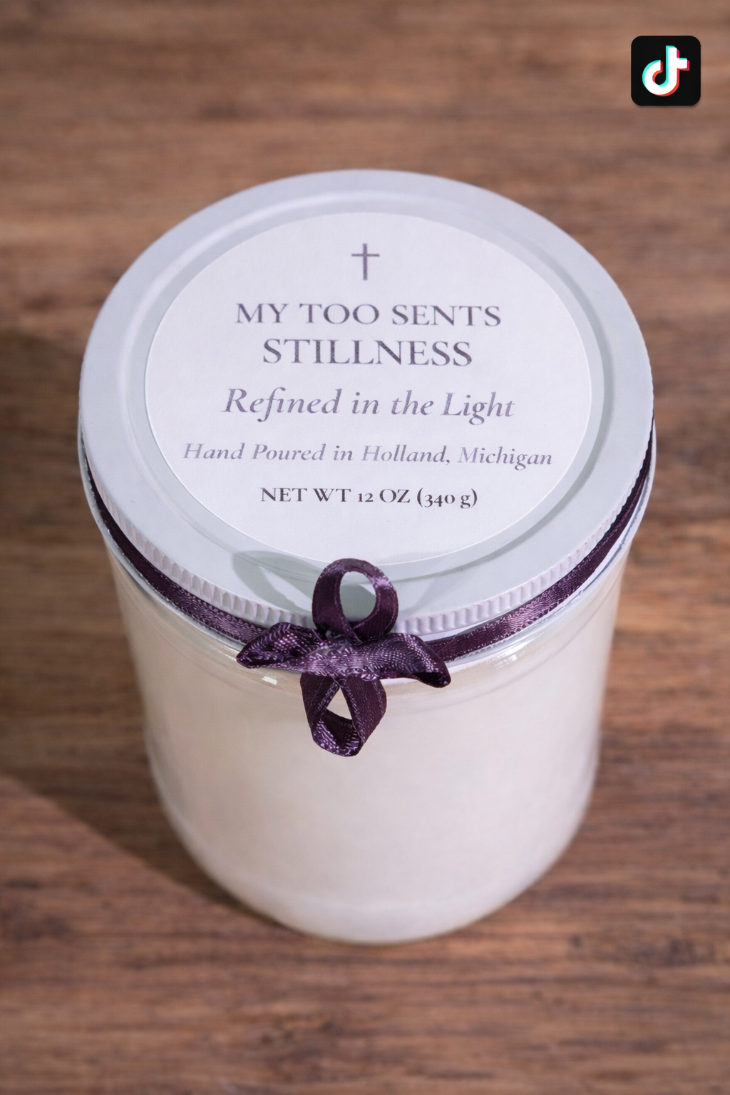 A candle in a glass jar with a white lid and a purple ribbon tied around the lid's edge. The lid has a label reading 'My Too Sents Stillness, Refined in the Light, Hand Poured in Holland, Michigan, Net Wt 12 Oz (340 g).' The candle is placed on a wooden surface.