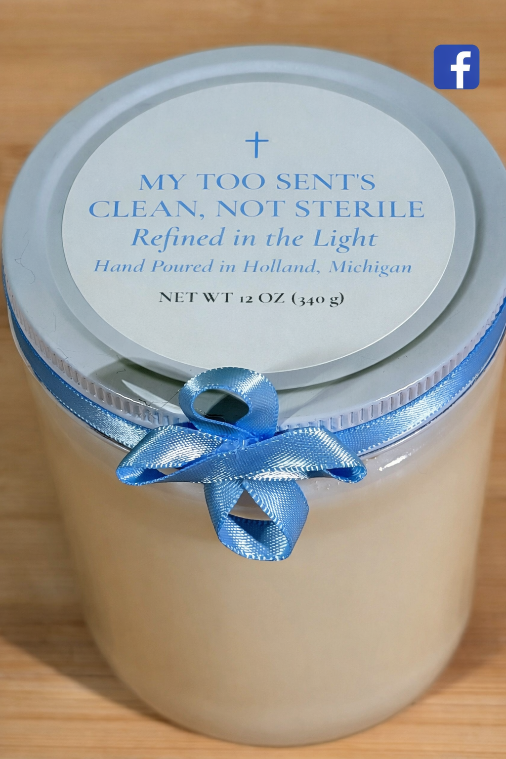 A container of scented candle with a light blue lid, tied with a matching blue satin ribbon, and a label that reads 'My Too Sent's Clean, Not Sterile, Refined in the Light, Hand Poured in Holland, Michigan, 12 oz.'