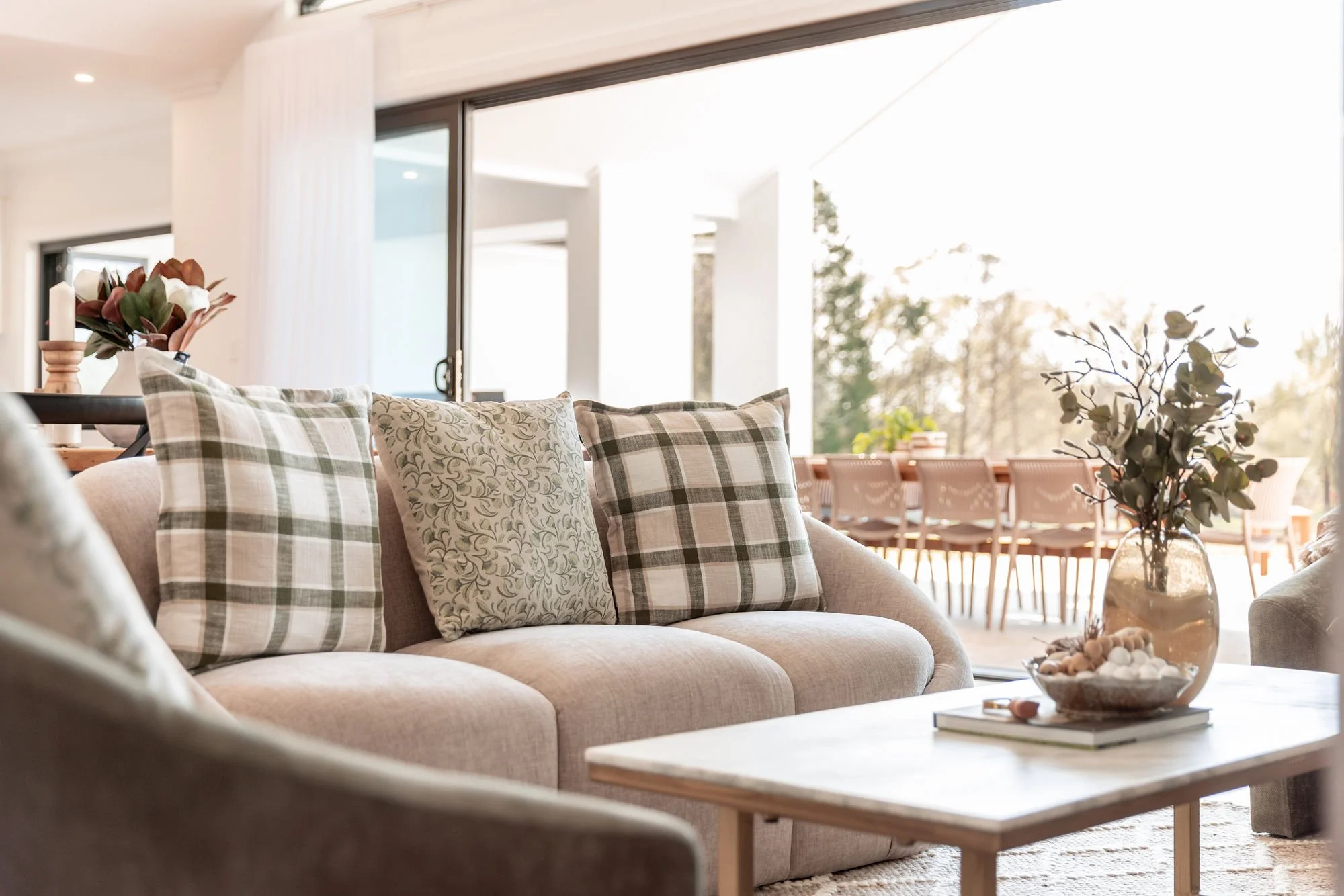 A cozy living room with a beige sofa adorned with five plaid and floral-patterned cushions, a coffee table with a vase of branches and a decorative bowl, and a large sliding glass door leading to an outdoor patio with a dining table and chairs, surro