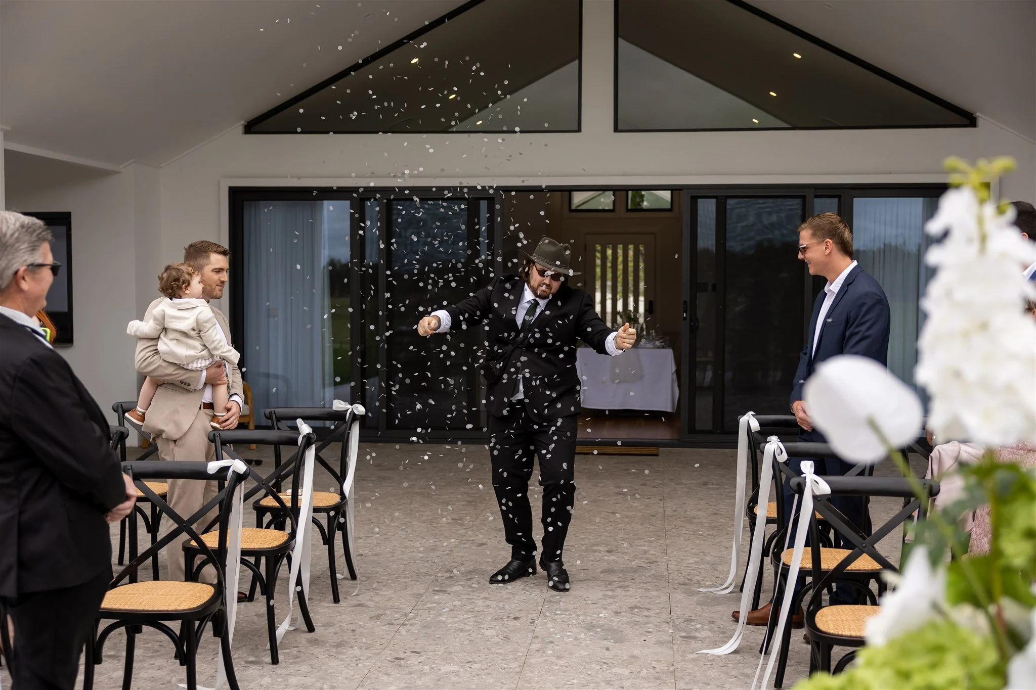 A man dressed in a black suit, hat, and sunglasses dancing among confetti at a wedding reception outside a modern house with sliding glass doors, with guests watching and smiling.