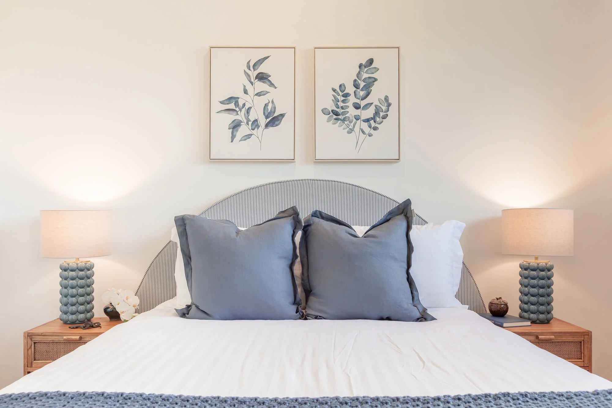 A neatly made bed with a gray and white color scheme, featuring a gray headboard, two large gray pillows, two white pillows, and a textured gray blanket at the foot. Two matching lamps with round, bead-like bases and beige lampshades flank the bed, w