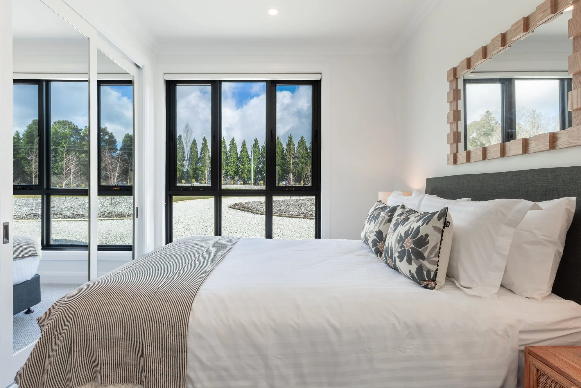 A bedroom with a large bed, decorative pillows, a wooden framed mirror, and large windows showing a view of trees and a cloudy sky outside.