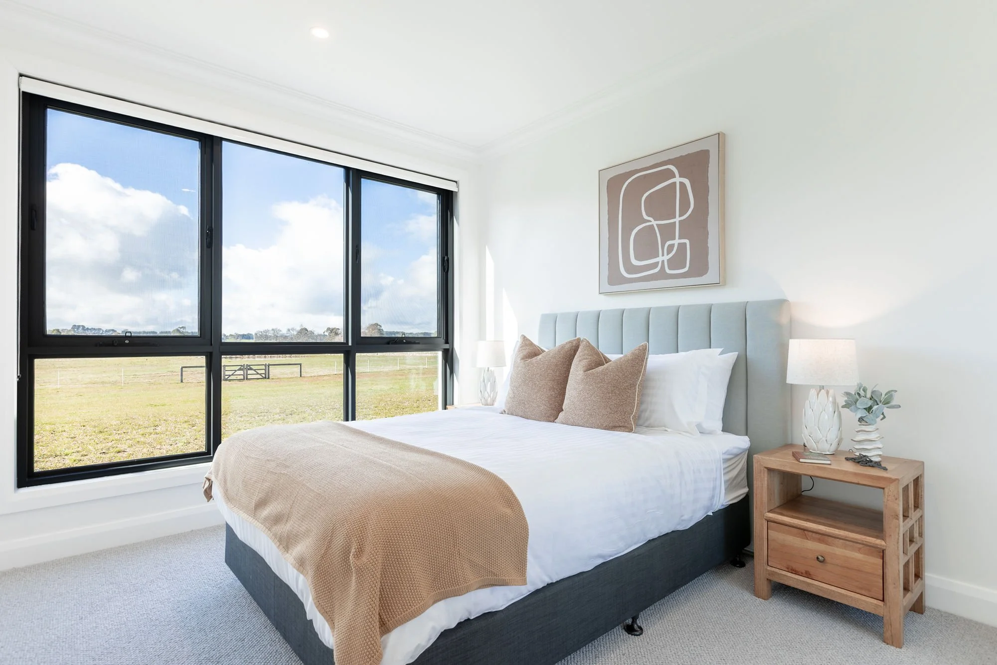 Bright bedroom with a large window showing a grassy field outside, a bed with white linens and beige pillows, a wooden nightstand with a lamp and decorative vase, modern abstract wall art.