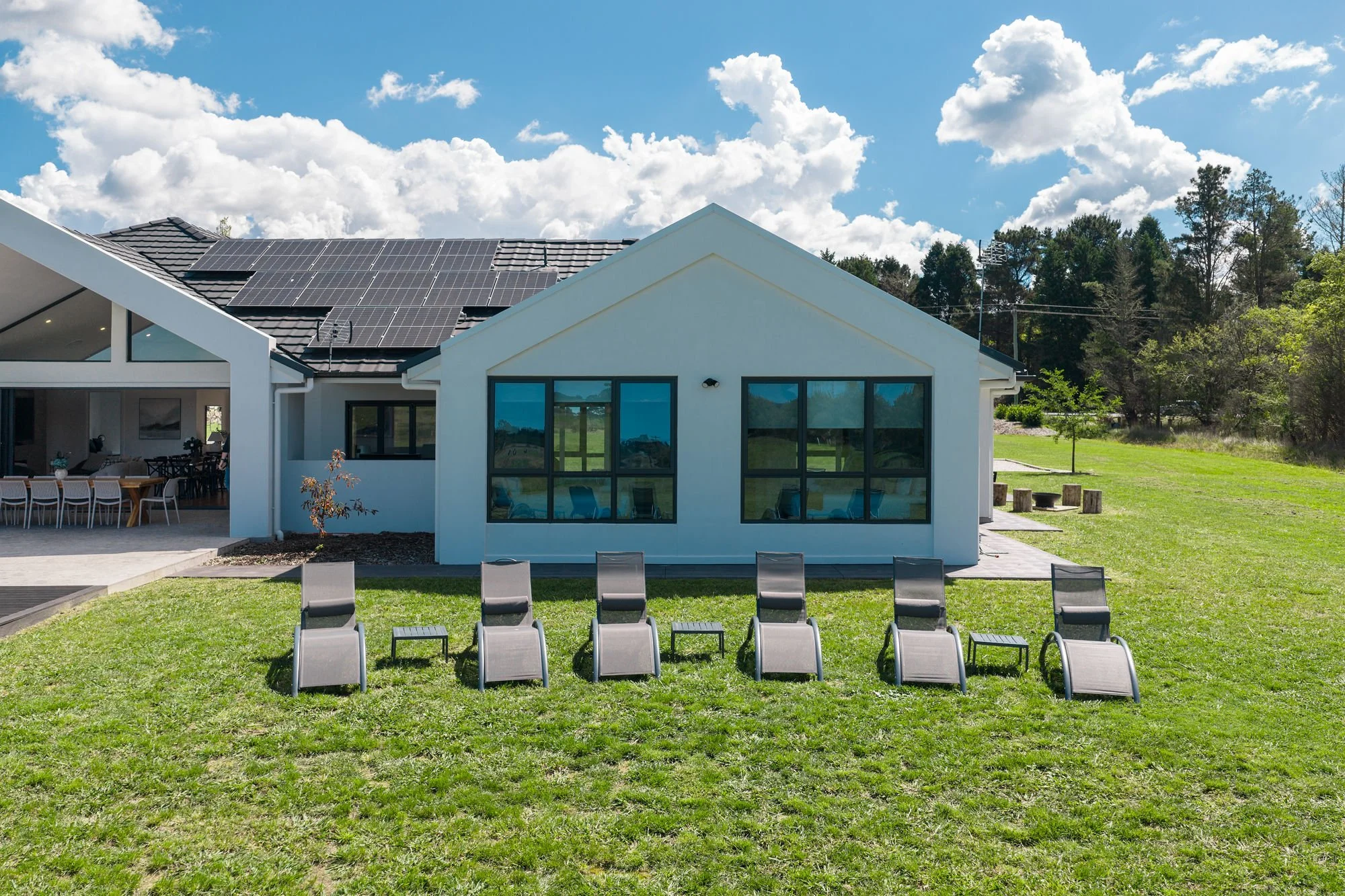Modern house with solar panels on roof, outdoor seating area, and eight lounge chairs on grassy lawn under partly cloudy sky.