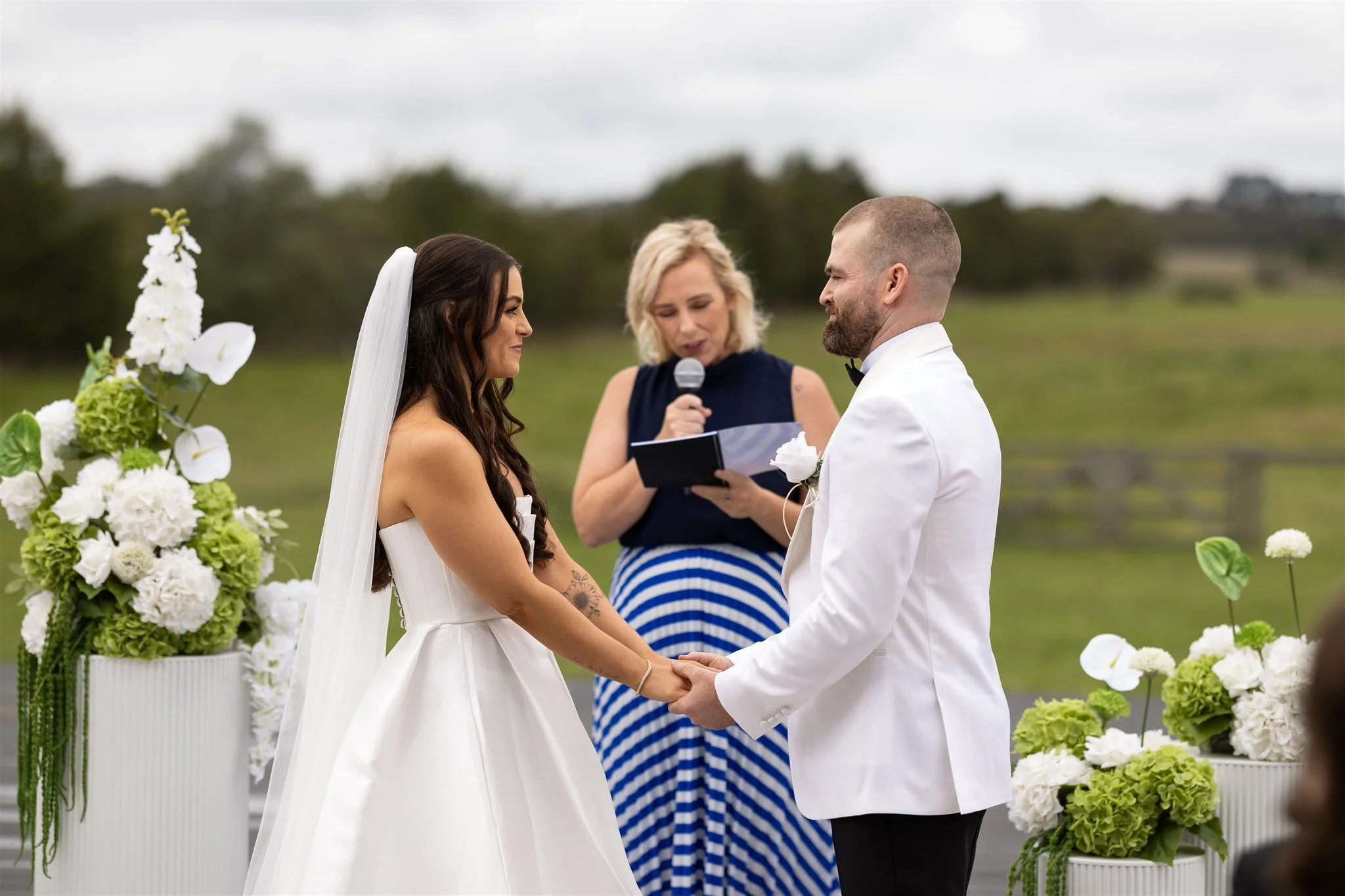A bride and groom holding hands during an outdoor wedding ceremony, with an officiant speaking into a microphone behind them. The bride is wearing a white wedding dress with a veil, and the groom is in a white tuxedo jacket. There are large flower ar