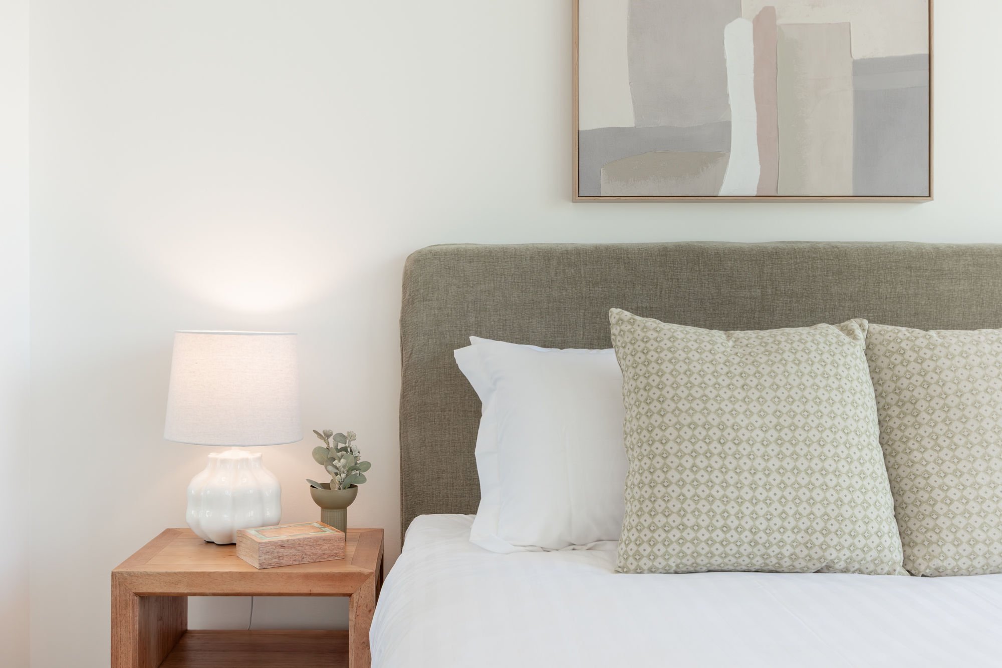 A cozy bedroom with a beige upholstered headboard, two patterned pillows, and white bedding. A wooden nightstand holds a white lamp and a small potted plant. Abstract artwork hangs above the bed on a white wall.