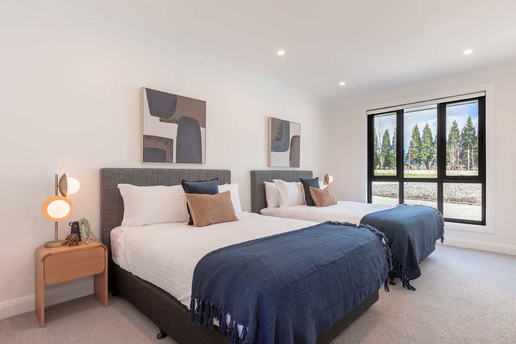 A bedroom with two beds, each with white bedding, dark blue blankets, and a mix of navy and beige pillows. The room has a large window showing a view of trees outside, modern art above each bed, and a wooden nightstand with a lamp and decor items.