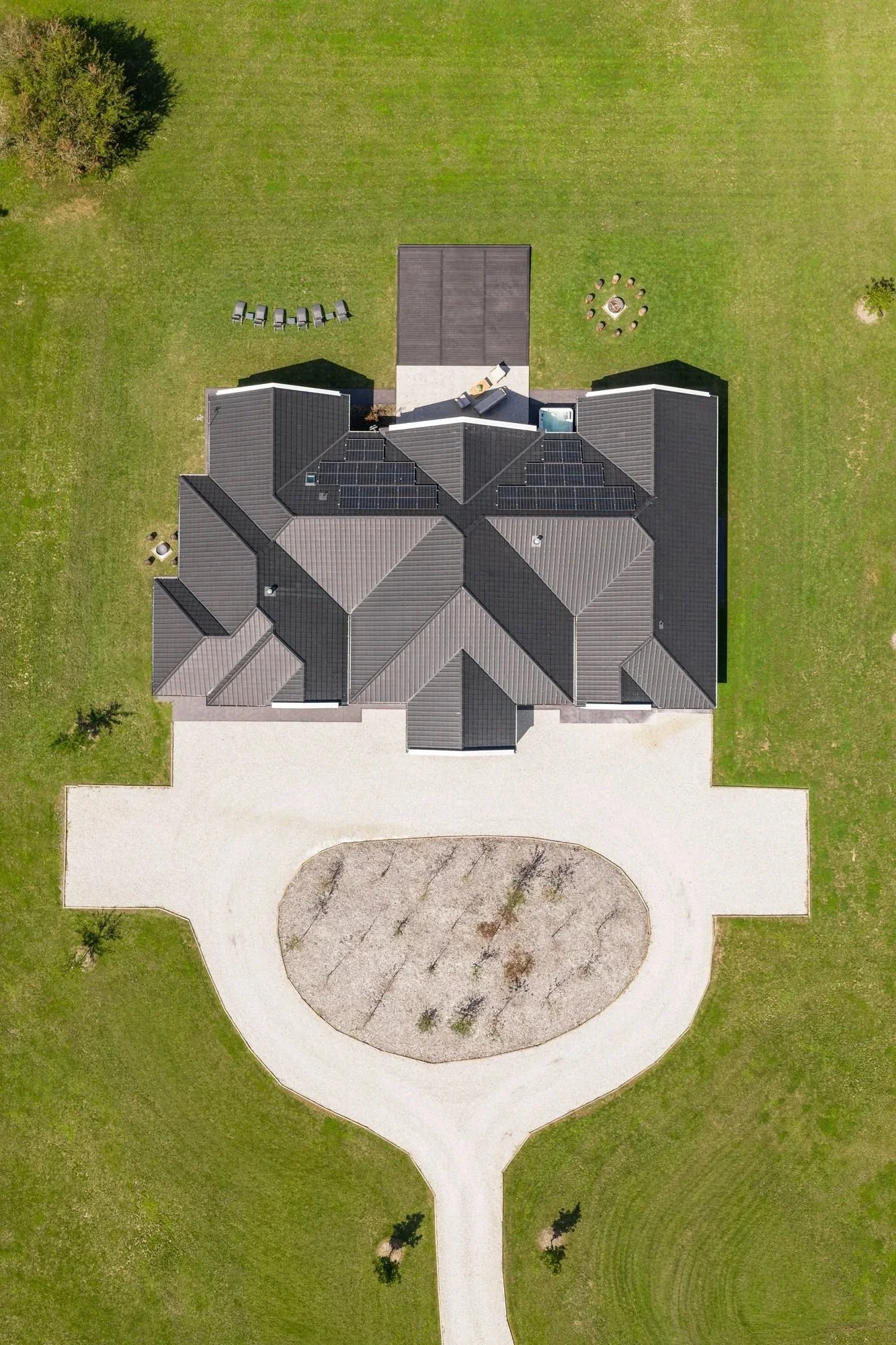 Aerial view of a large house with a gray roof, white driveway, and green lawn. The house features solar panels on the roof and a backyard with a fire pit surrounded by chairs and a small deck with a hot tub.