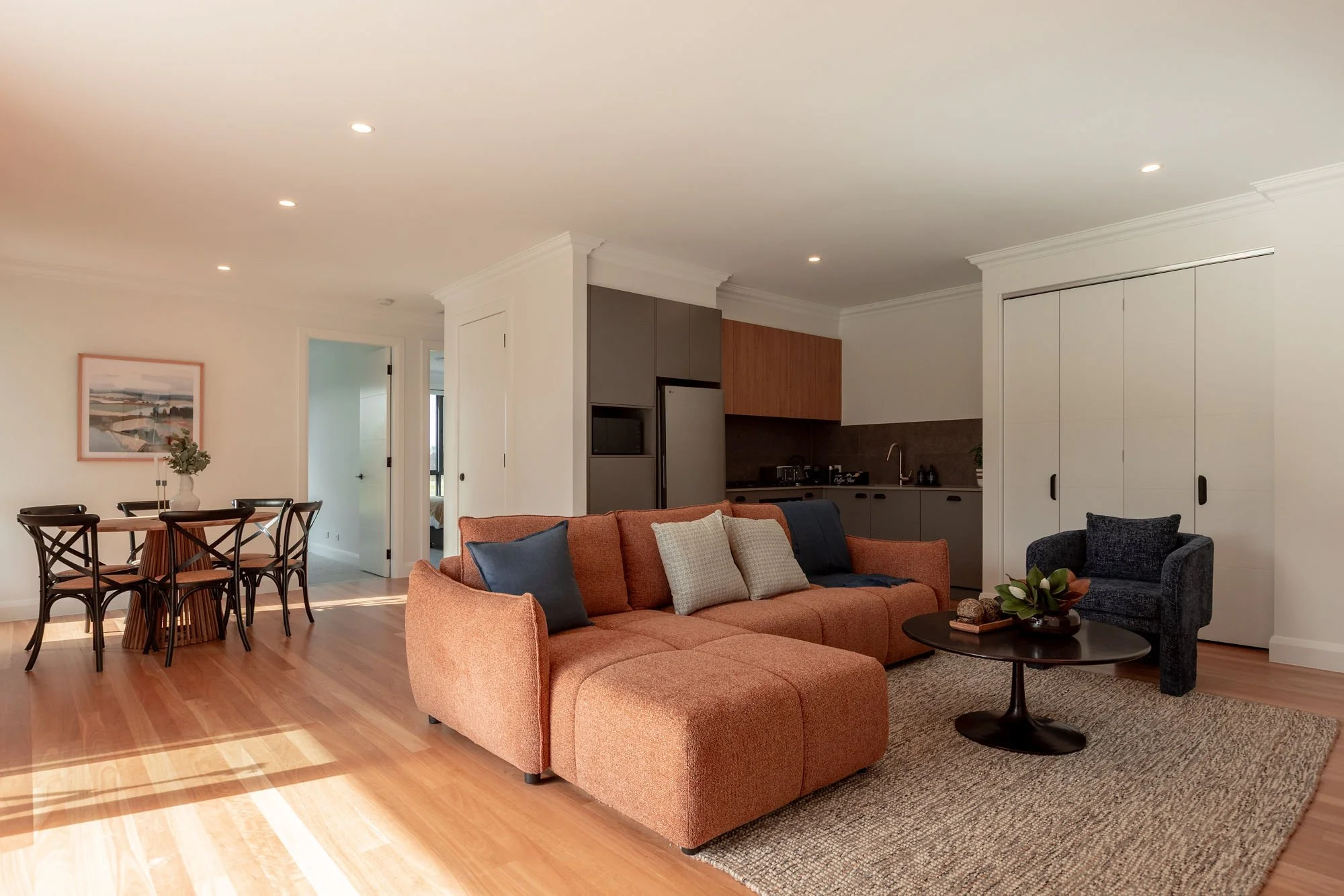 Living room with a coral sectional sofa, a dark blue armchair, a round black coffee table with a vase and plants, a beige textured rug, and a dining table with black chairs in the background, with kitchen area and a gallery wall in the distance.