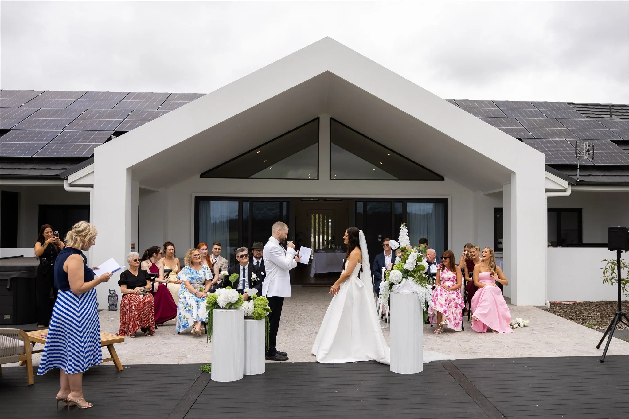 A couple gets married outdoors with guests sitting on chairs, in front of a modern white house with solar panels on the roof, on a cloudy day.