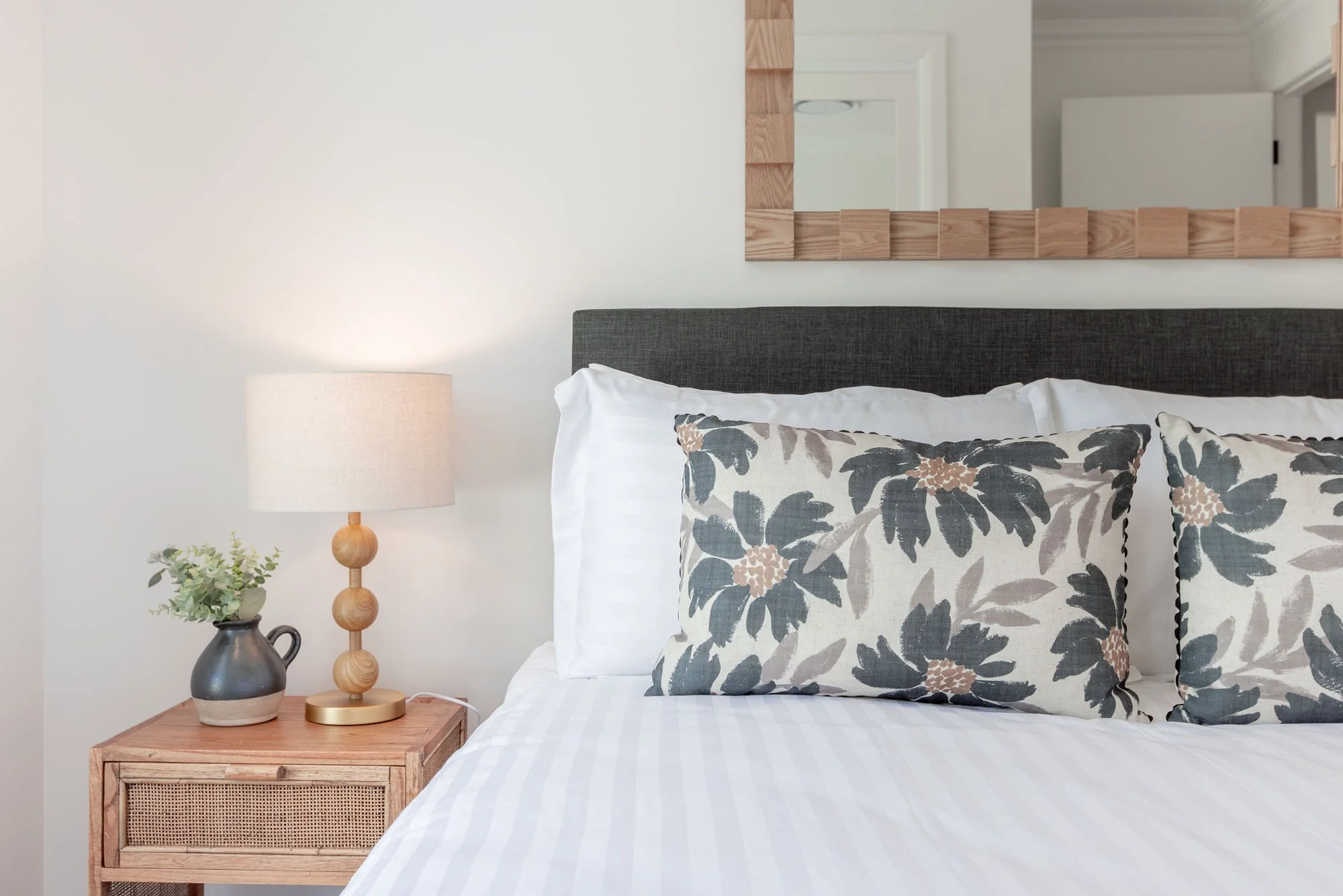 A bedroom with a dark gray headboard, floral pillows, a white bedside table, a beige lamp, and a small plant. A mirror hangs above the bed.