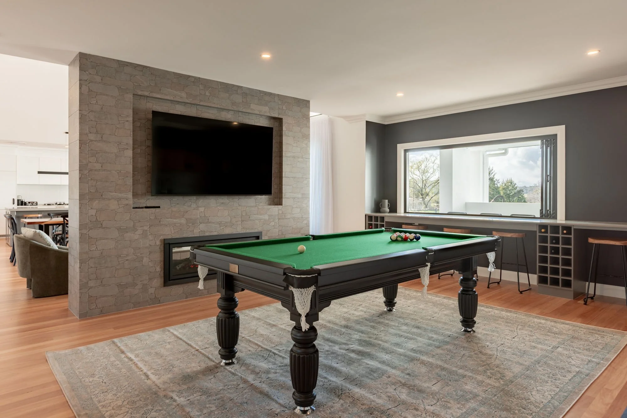 A modern living room featuring a black pool table with green felt, pool balls, and a cue ball, situated on a patterned rug. There's a large window with a view of trees, a stone fireplace with a mounted flat-screen TV, and a grey wall with bar stools 