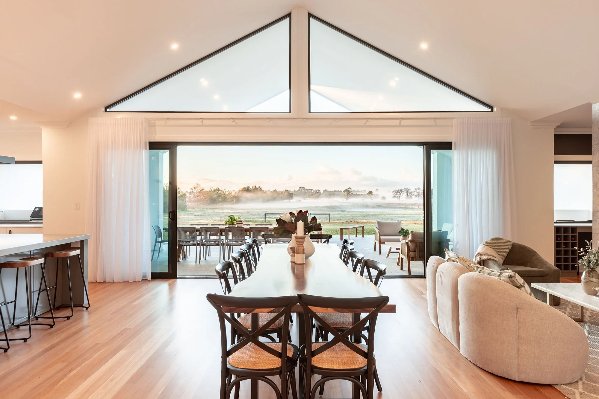 Bright, open living and dining area with large glass sliding doors leading to an outdoor patio overlooking a foggy landscape with trees and a field, featuring a long dining table with black chairs, a cozy sitting area with armchairs and a sofa, and modern decor.
