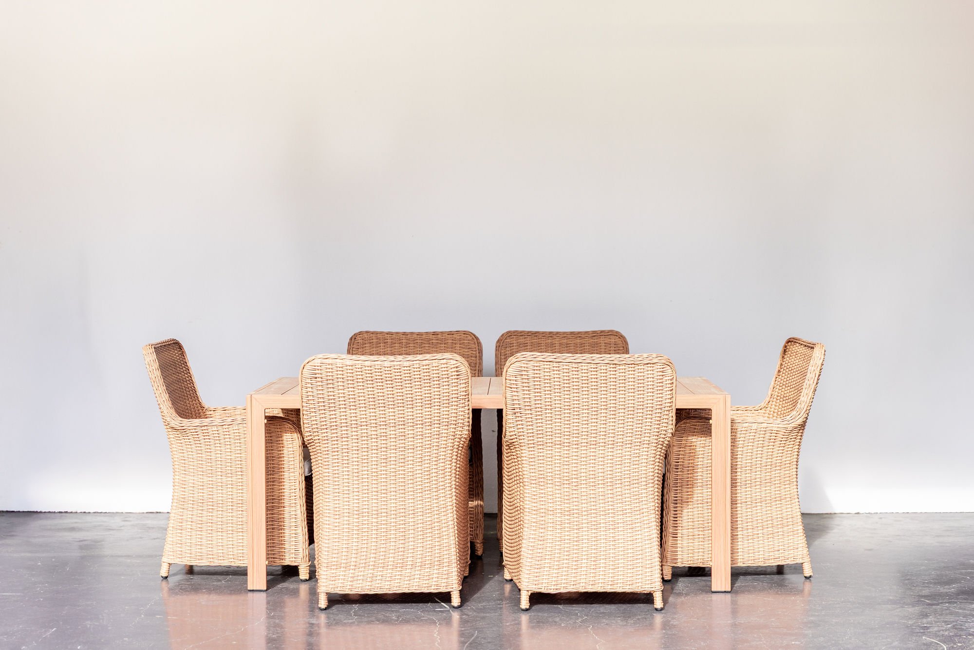 A dining table with six wicker chairs arranged around it, set against a plain white wall.
