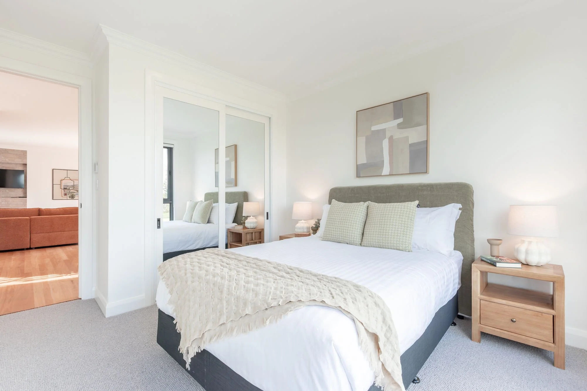 Bedroom with a bed having a green headboard, white bedding, and pillows, beside a wooden nightstand with a lamp and books. A mirror sliding closet door reflects part of the room, and there is artwork above the bed. Past the bedroom door, a living roo