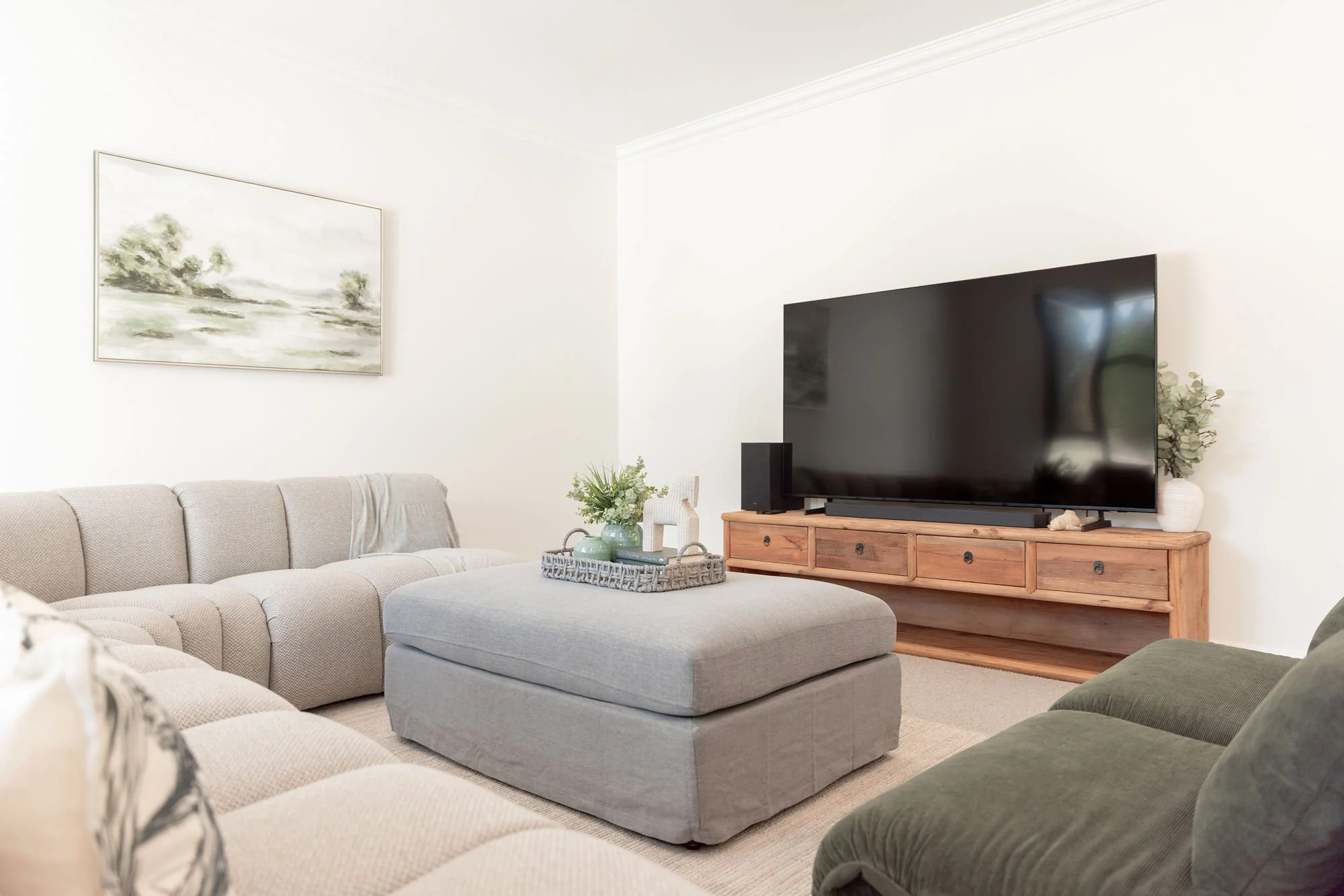 Living room with a beige sectional sofa, a gray ottoman with a tray of decorative items, a wooden TV stand with a large flat-screen TV, a green armchair, and a framed landscape painting on the wall.