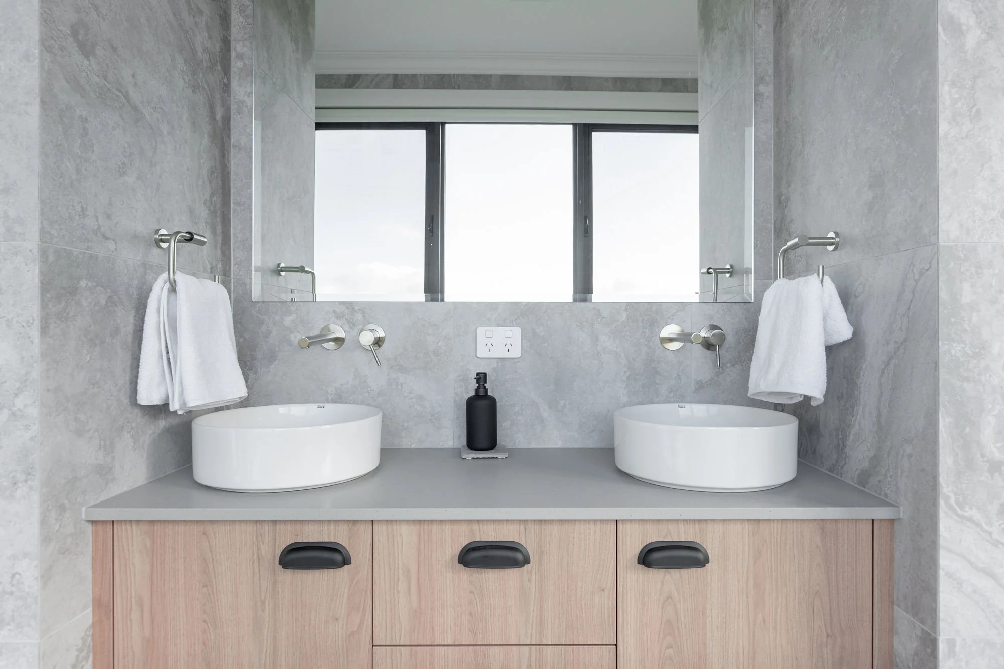Modern bathroom with two white vessel sinks, large mirror, and gray marble walls and countertops. Two white towels hang on silver towel bars, and black soap dispenser sits between the sinks. There is a large window in the reflection of the mirror.