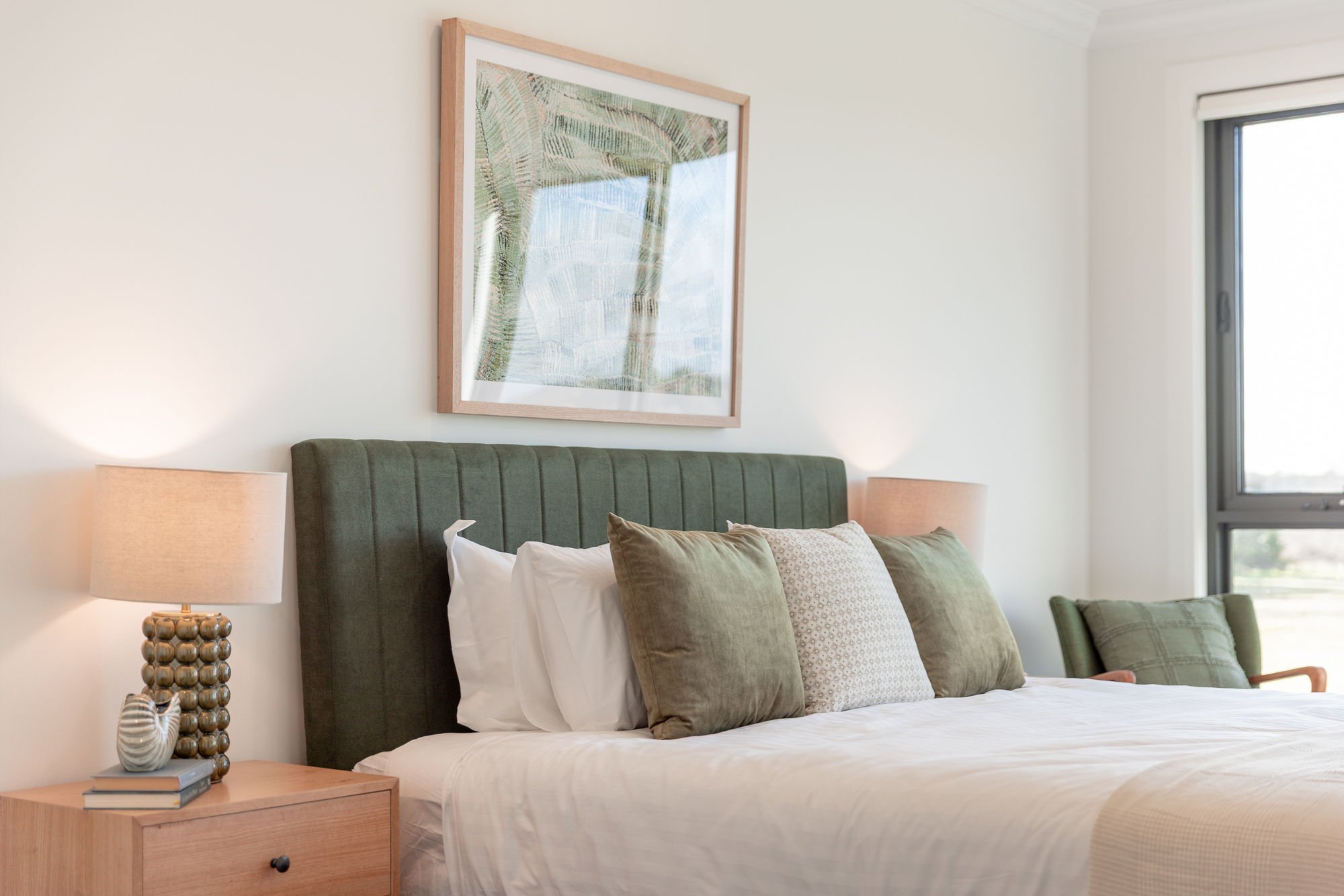 A bedroom with a bed, green upholstered headboard, and multiple pillows. There are wooden nightstands with lamps, a framed artwork above the bed, and a window with a view outside.