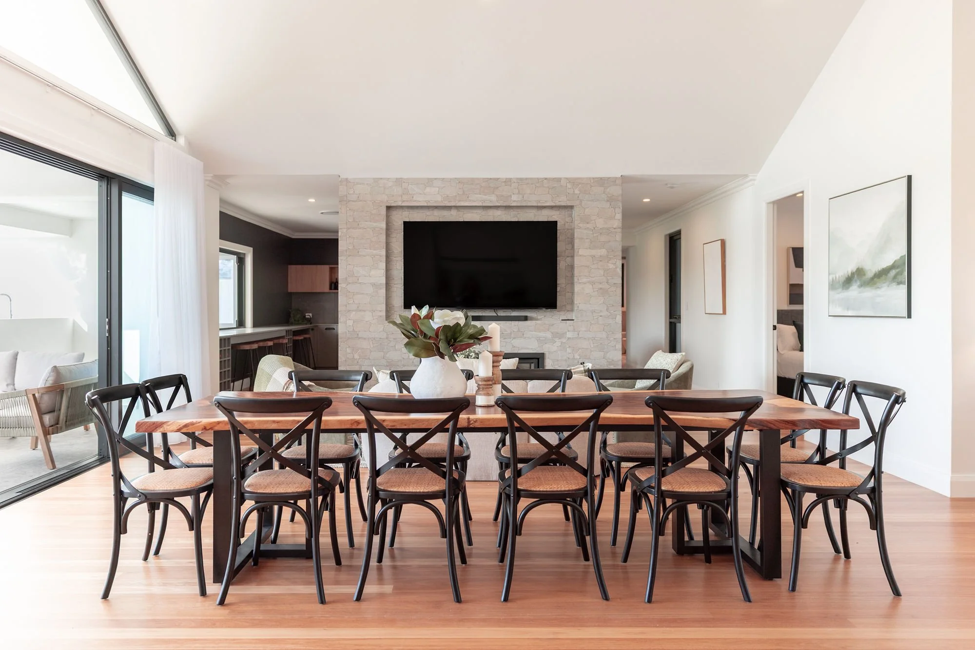 Spacious dining area with a wooden table, black chairs, a white vase with greenery, located in a modern home with a large TV on a stone wall, and artwork on the white walls.
