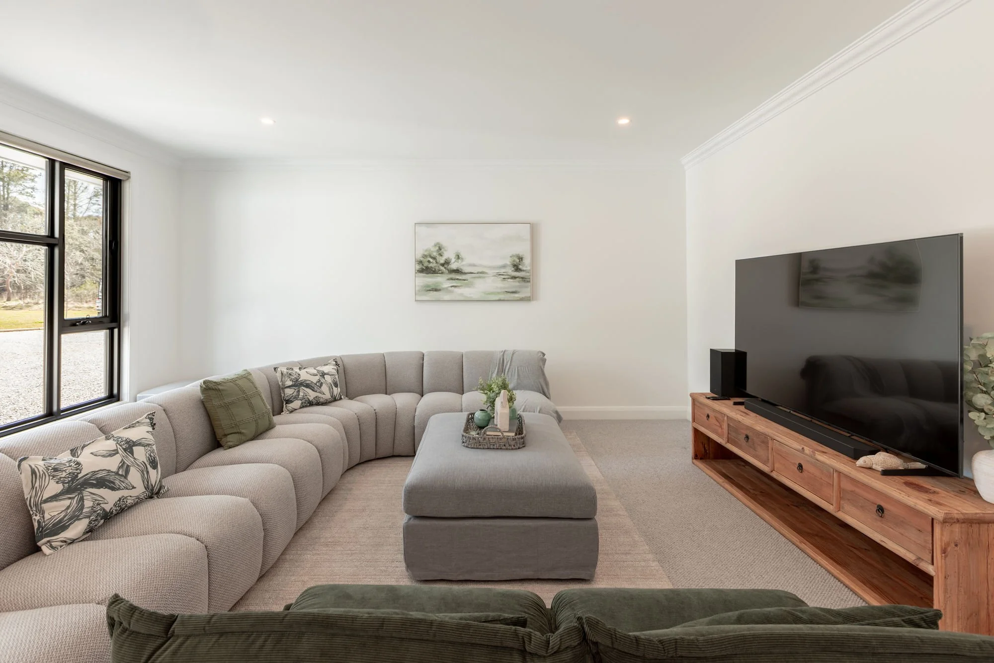 Living room with a large light gray sectional sofa, decorative pillows, a matching ottoman with a tray and decorative items, a wooden TV stand with a large flat-screen TV, window on the left letting in natural light, a wall art picture, and a carpete