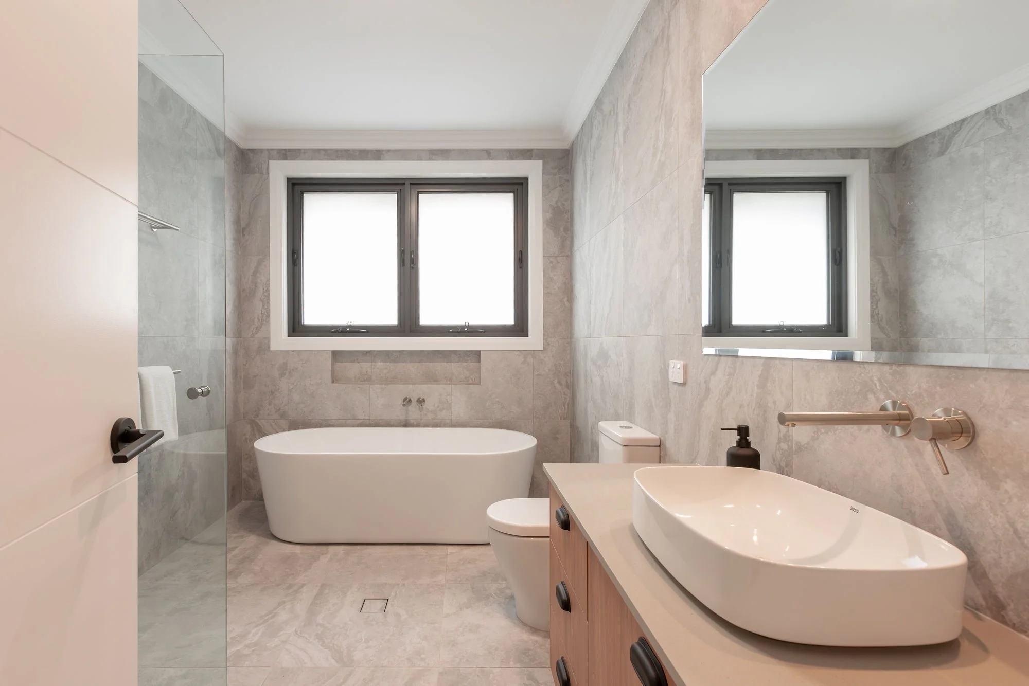 Modern bathroom with a bathtub, toilet, sink, large mirror, and two windows with frosted glass.