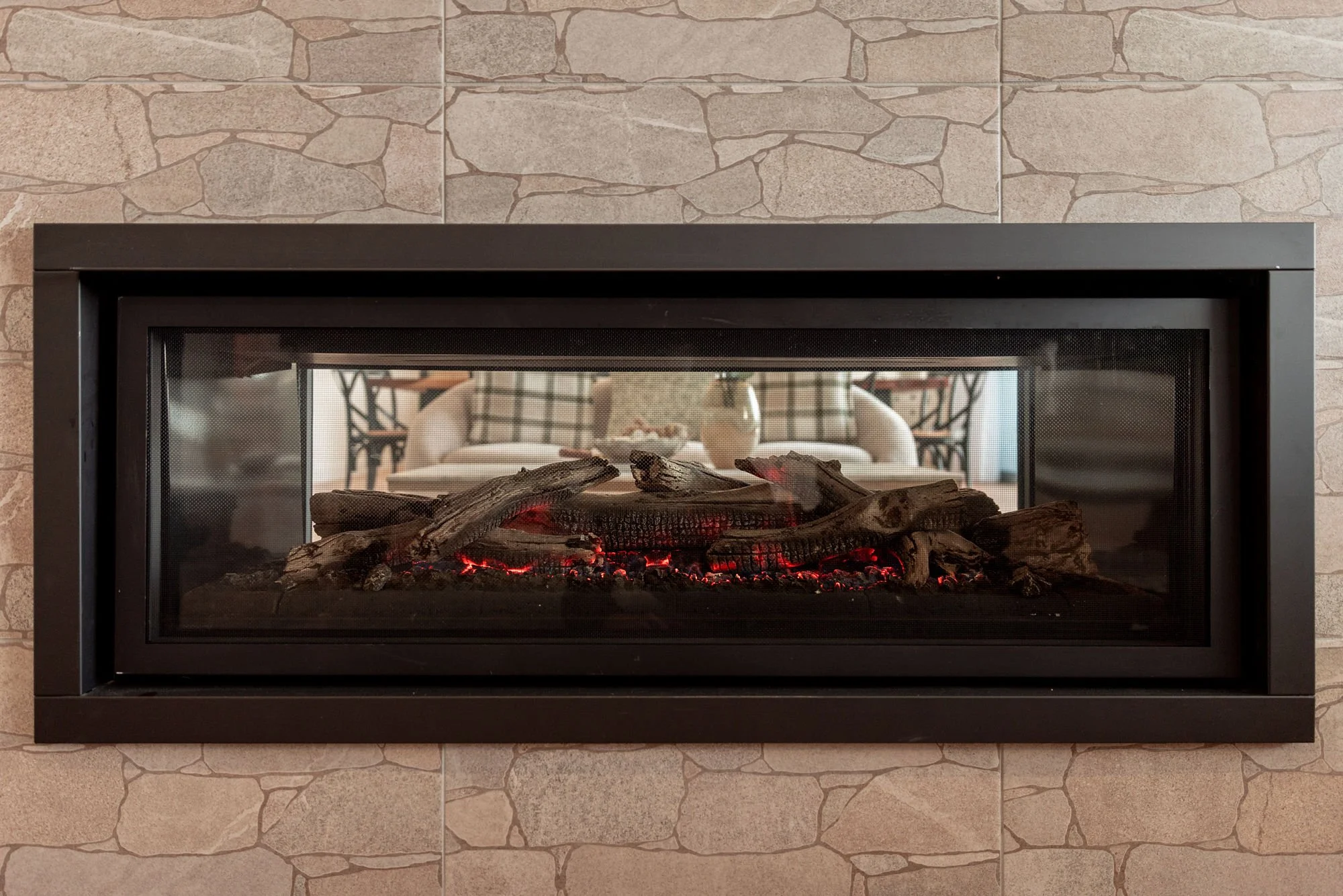 Indoor fireplace with black frame and glass front, filled with glowing artificial logs, mounted on a stone wall