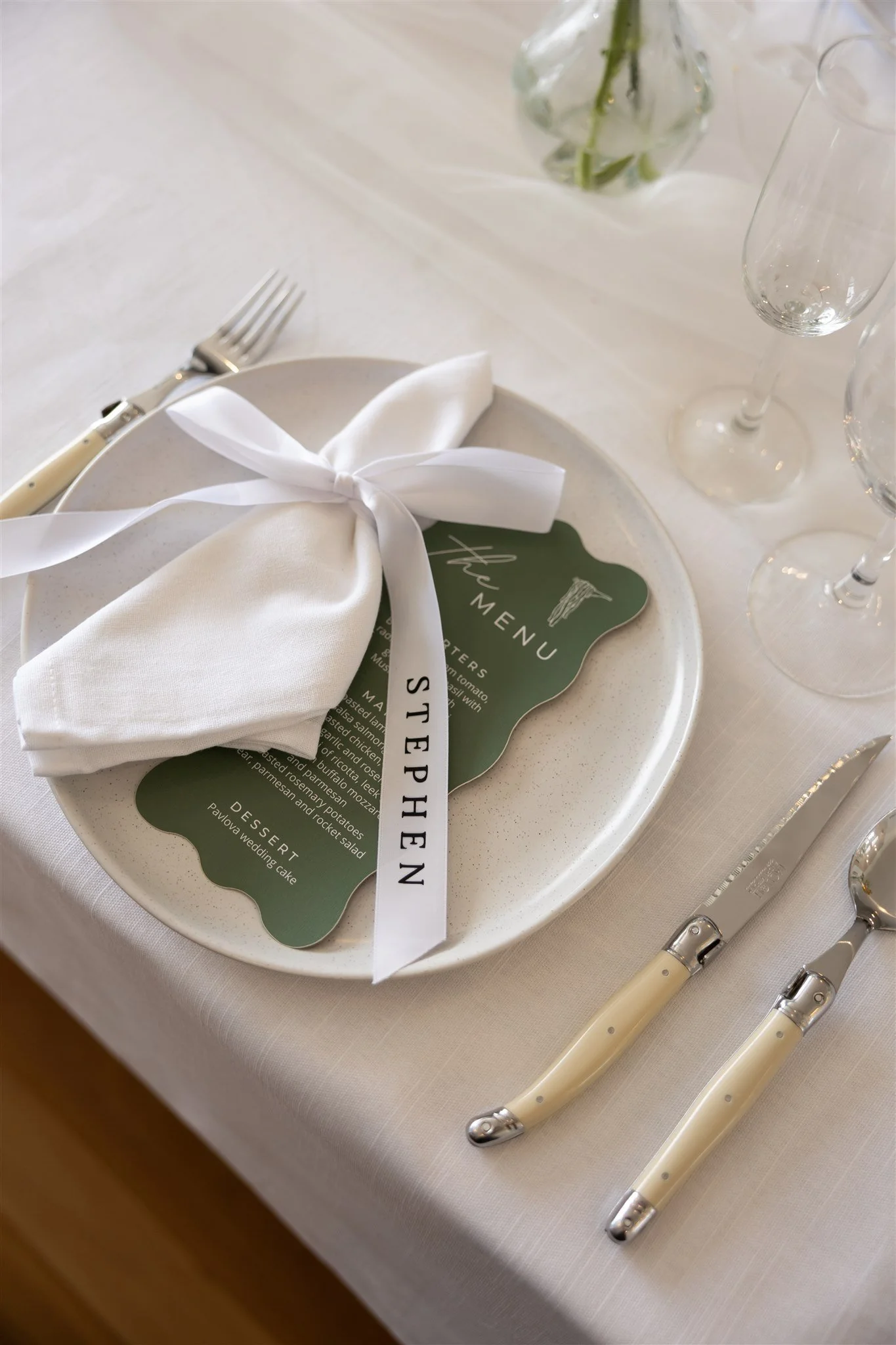 Elegant table setting with a white plate, a white cloth napkin tied with a white ribbon labeled "Stephen," silverware, wine glasses, and a green menu on the plate. A small vase with flowers is in the background.