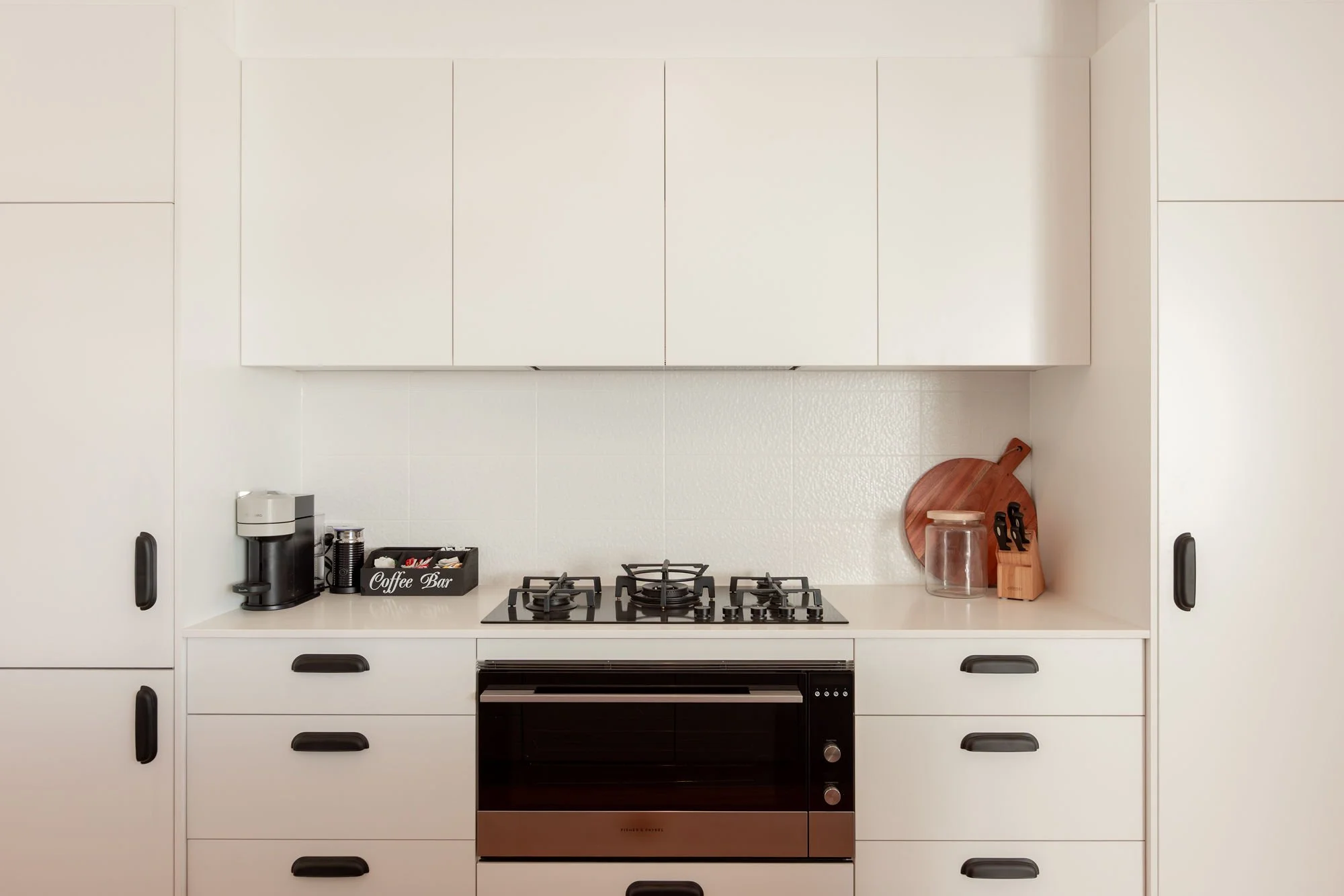 Minimalist kitchen with white cabinets, a built-in oven, a gas stove, and countertop accessories including a coffee maker, a tray labeled 'Coffee Bar', a glass jar, a wooden cutting board, and a knife block.