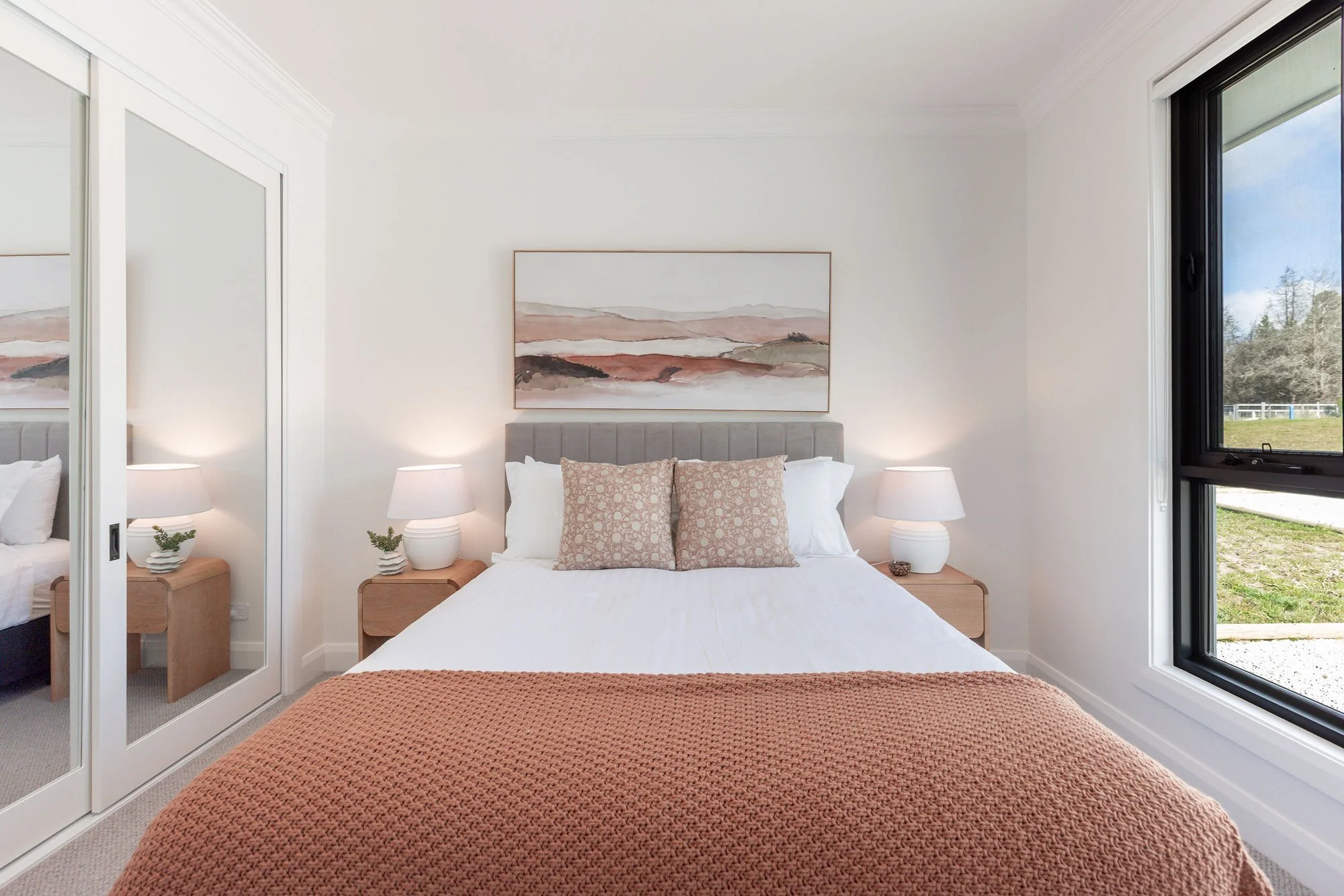 A bedroom with a bed, two nightstands with lamps, a window showing a green outdoor area, a framed landscape painting above the headboard, and a mirrored closet door reflecting part of the room.