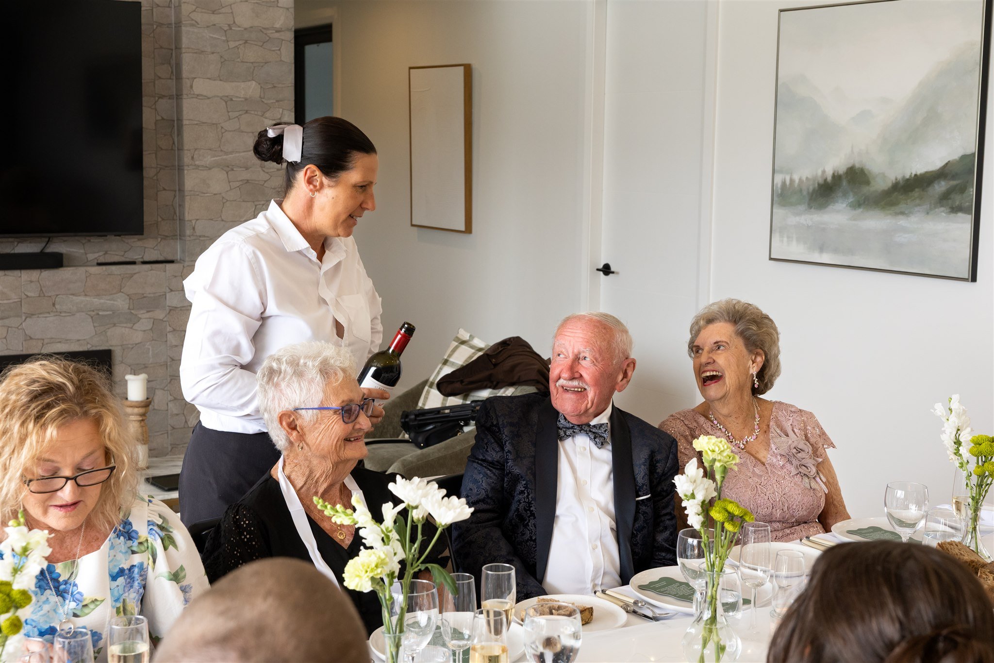 Group of elderly people at a dinner table with a woman serving wine, smiling and engaging in conversation, with floral decorations and wine glasses on the table.