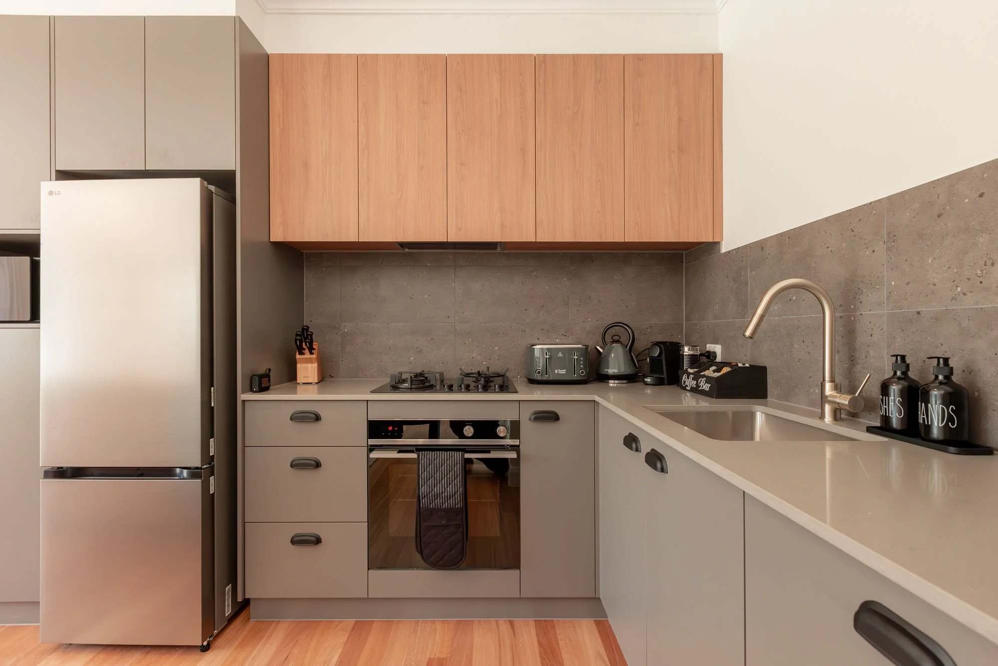 Modern kitchen with gray lower cabinets, wooden upper cabinets, stainless steel refrigerator, countertop appliances including toaster, kettle, and coffee maker, a sink with a brass faucet, and labeled soap and hand lotion bottles.