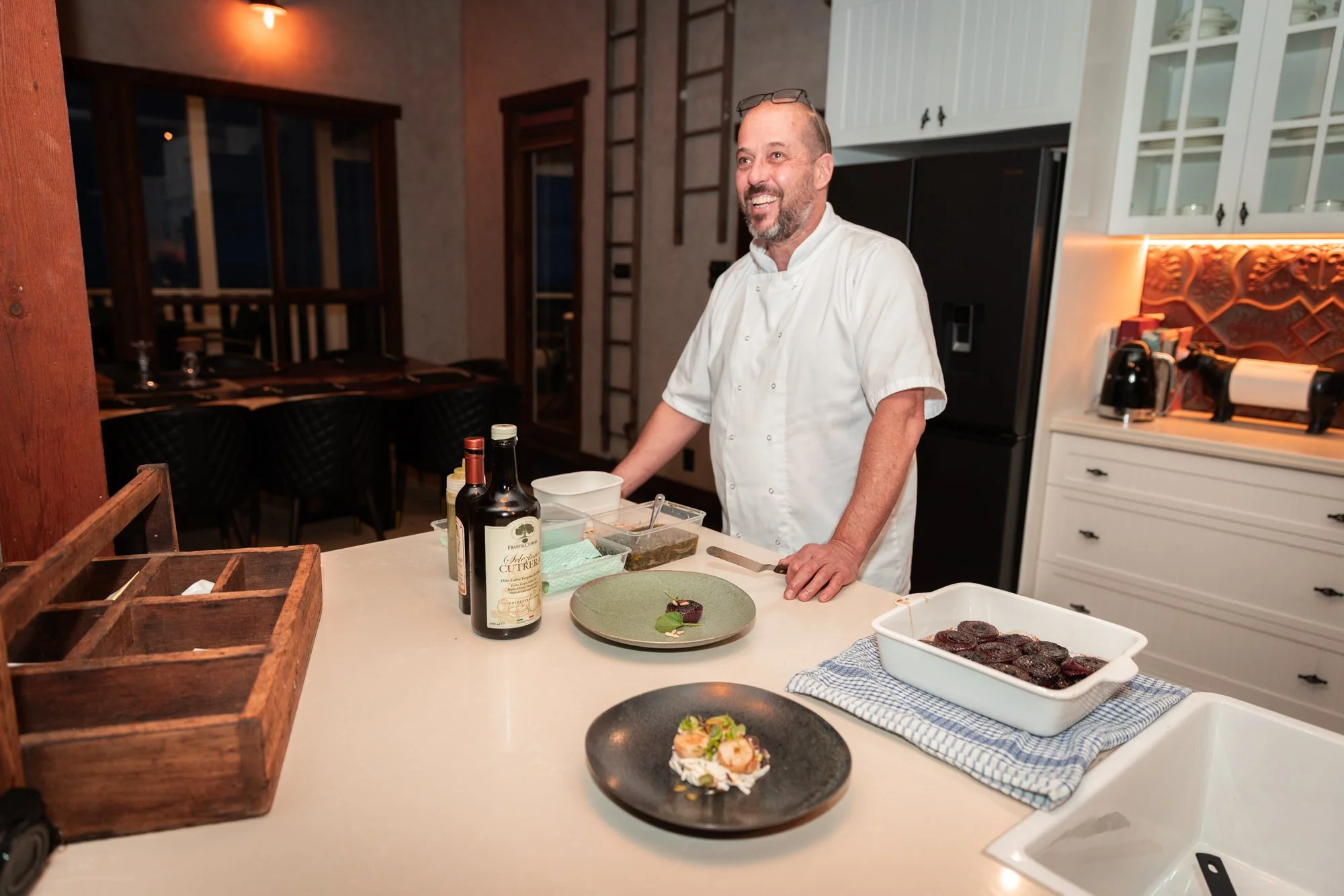 A smiling chef in a white uniform stands behind a kitchen counter with plated dishes and bottles of oil. The background features a well-lit kitchen and dining area with windows.
