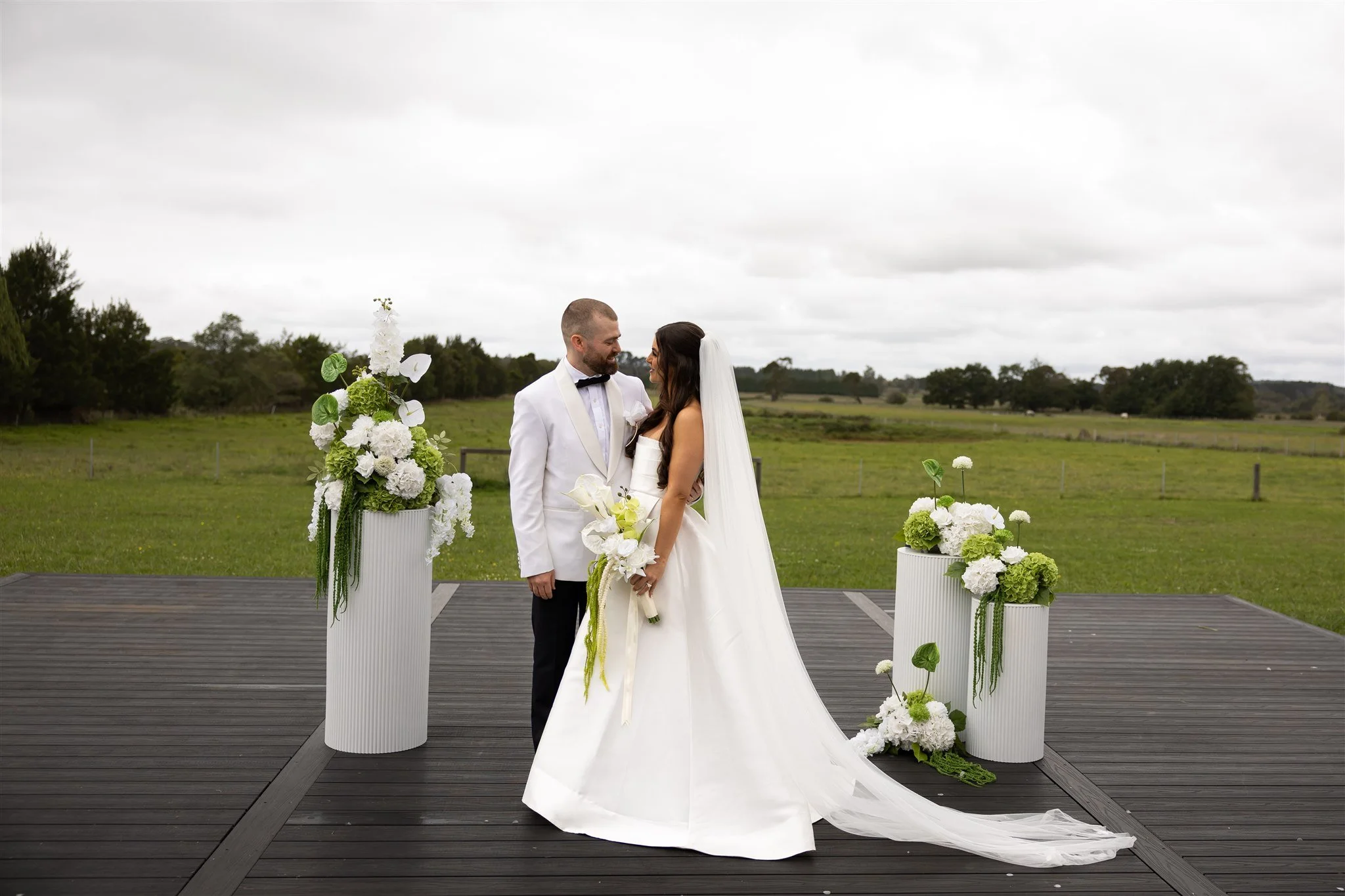 A bride and groom standing on a wooden platform, facing each other, with a field and trees in the background, decorated with tall white floral arrangements. The bride wears a white wedding gown with a long train and veil, holding a bouquet of white f
