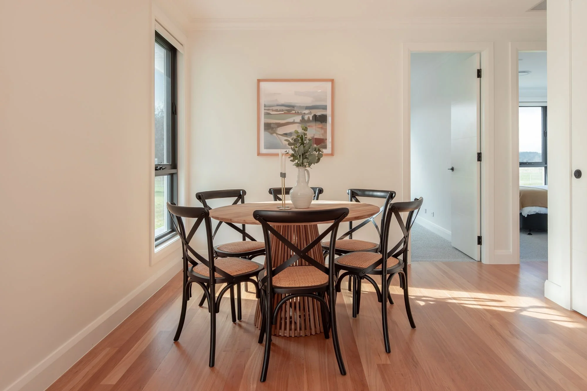 A dining room with a round wooden table surrounded by eight black chairs with woven seats. On the table is a white vase with green leaves and two candlesticks. The room has white walls, hardwood floors, and two windows letting in natural light. A fra