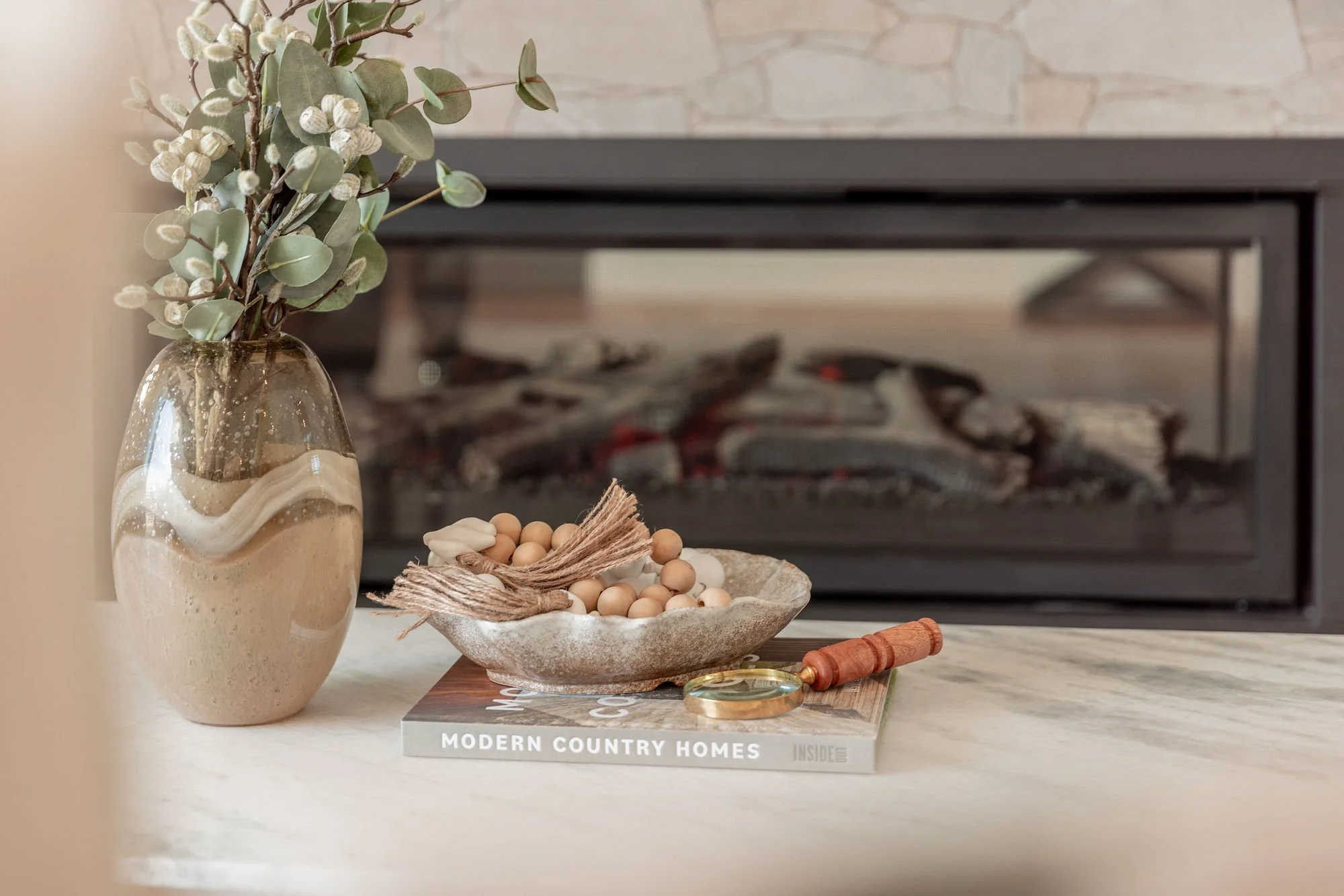 Decorative arrangement on a table with a vase of eucalyptus, a bowl of beads and crystals, a book titled 'Modern Country Homes,' a magnifying glass, and a small wooden rolling pin, with a fireplace in the background.