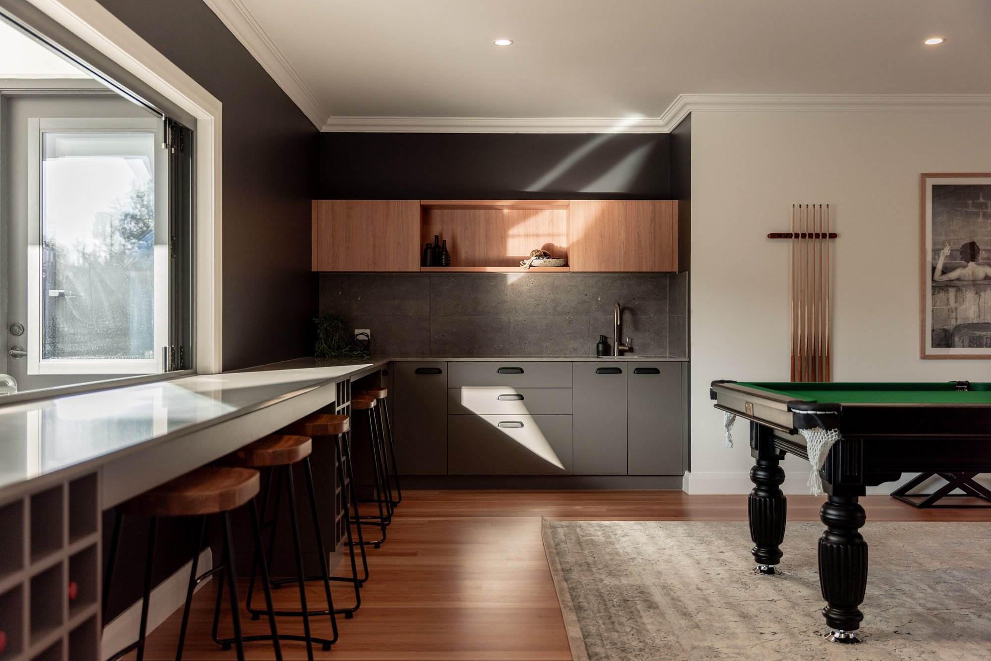Modern kitchen with gray lower cabinets, wooden upper cabinets, a dark gray backsplash, a sitting area with bar stools, a pool table, and a large window with sunlight streaming in.