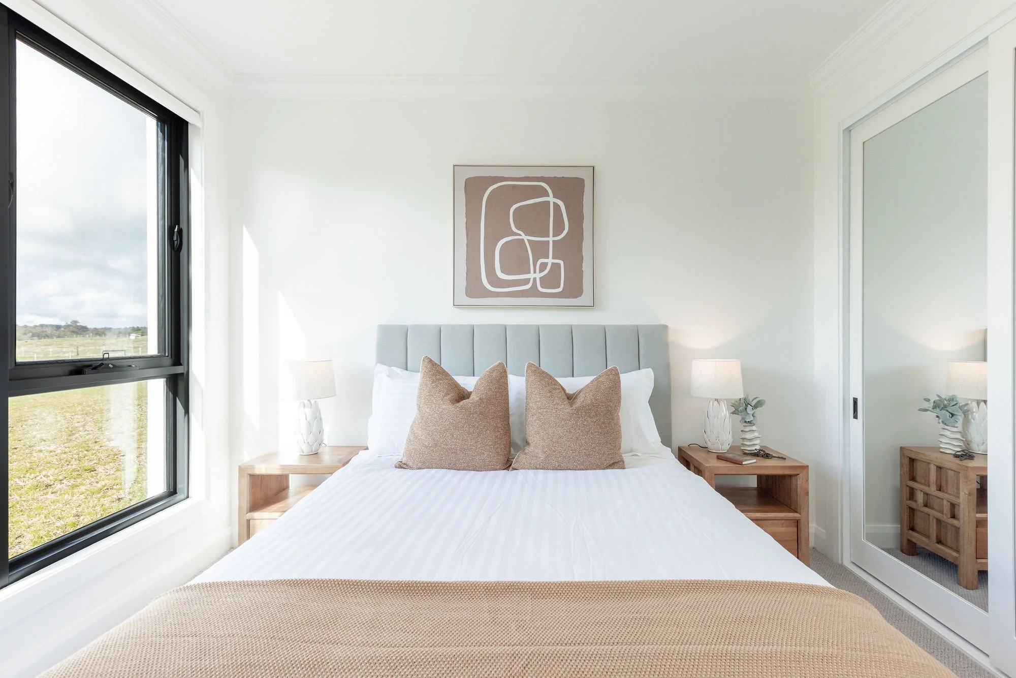A bedroom with a large bed, two wooden nightstands with lamps, beige pillows, and a window showing an outdoor landscape.
