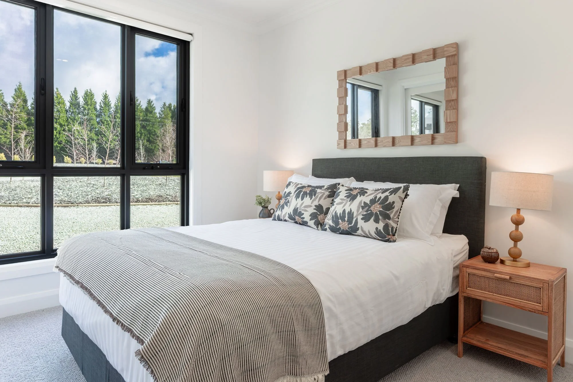A modern bedroom featuring a large bed with white bedding, decorative pillows, wooden side tables with lamps, a mirror above the bed, and large windows showing trees outside.